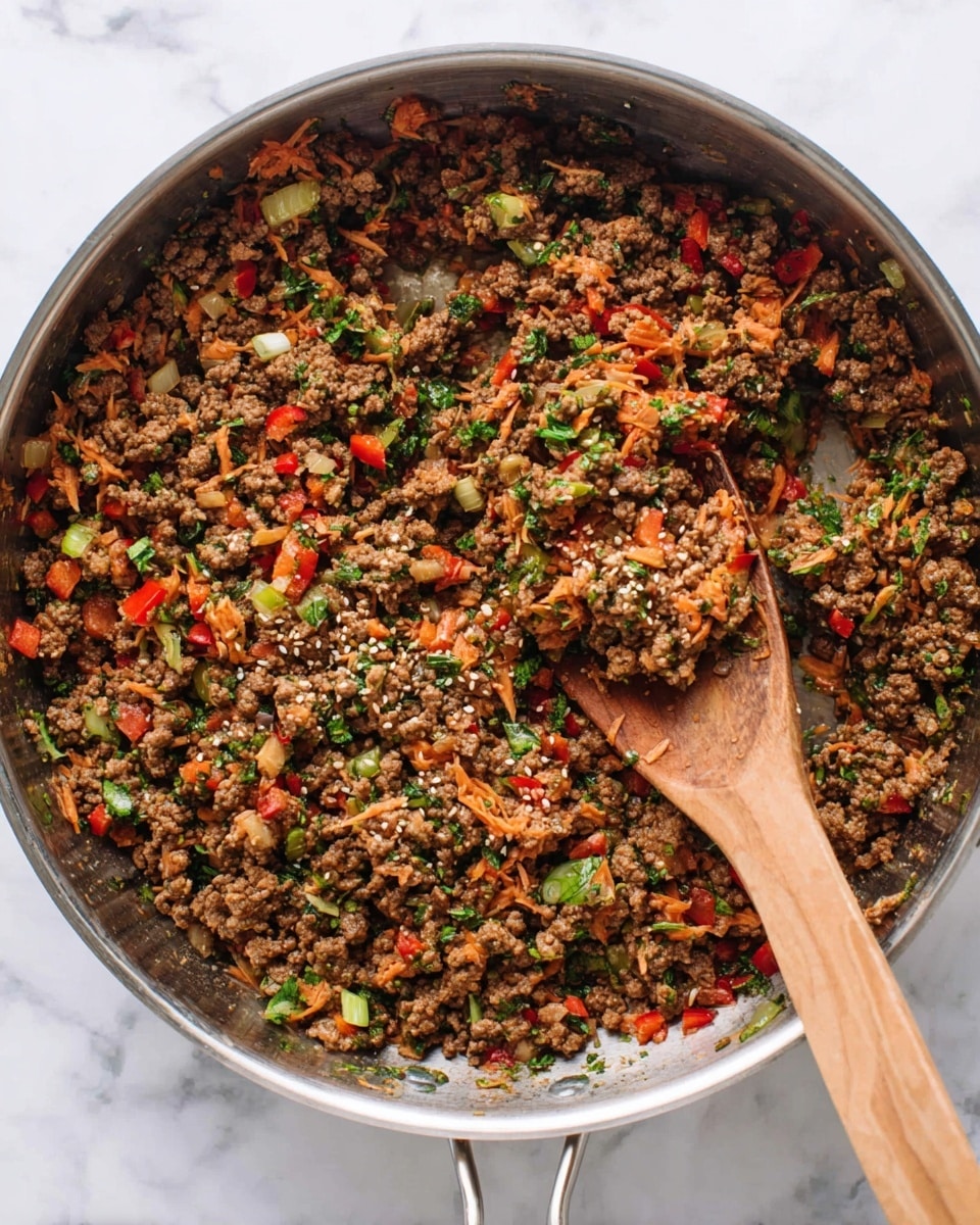 A large round silver pan is filled with cooked ground beef mixed with chopped vegetables including red bell peppers, onions, green onions, shredded carrots, and herbs scattered throughout. The beef is brown and crumbly, while the vegetables add pops of red, orange, and green colors. The mixture looks moist but not wet, with a few sesame seeds sprinkled on top. A wooden spoon rests inside the pan, partially covered in the food. The pan is set on a white marbled surface. Photo taken with an iphone --ar 4:5 --v 7