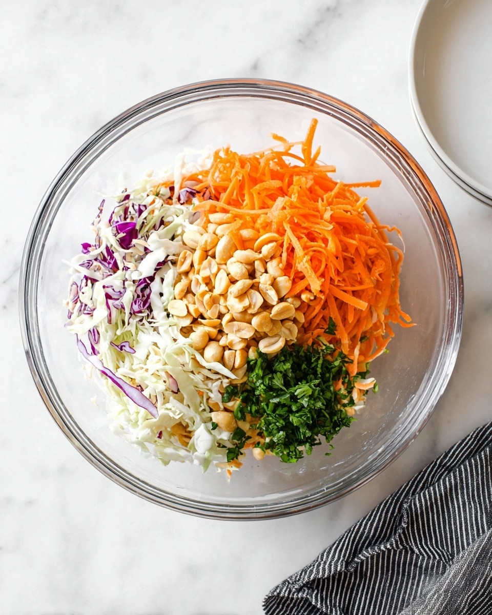 A clear glass bowl sitting on a white marbled surface holds four separate layers of salad ingredients: at the bottom is a mix of shredded white and purple cabbage with thin carrot strips, to the right is a heap of bright orange shredded carrots, in the center are whole light brown peanuts, and to the left is a small mound of finely chopped green herbs; a white plate is partially visible in the top right corner, and a black and white striped cloth rests at the bottom right corner of the image; photo taken with an iphone --ar 4:5 --v 7