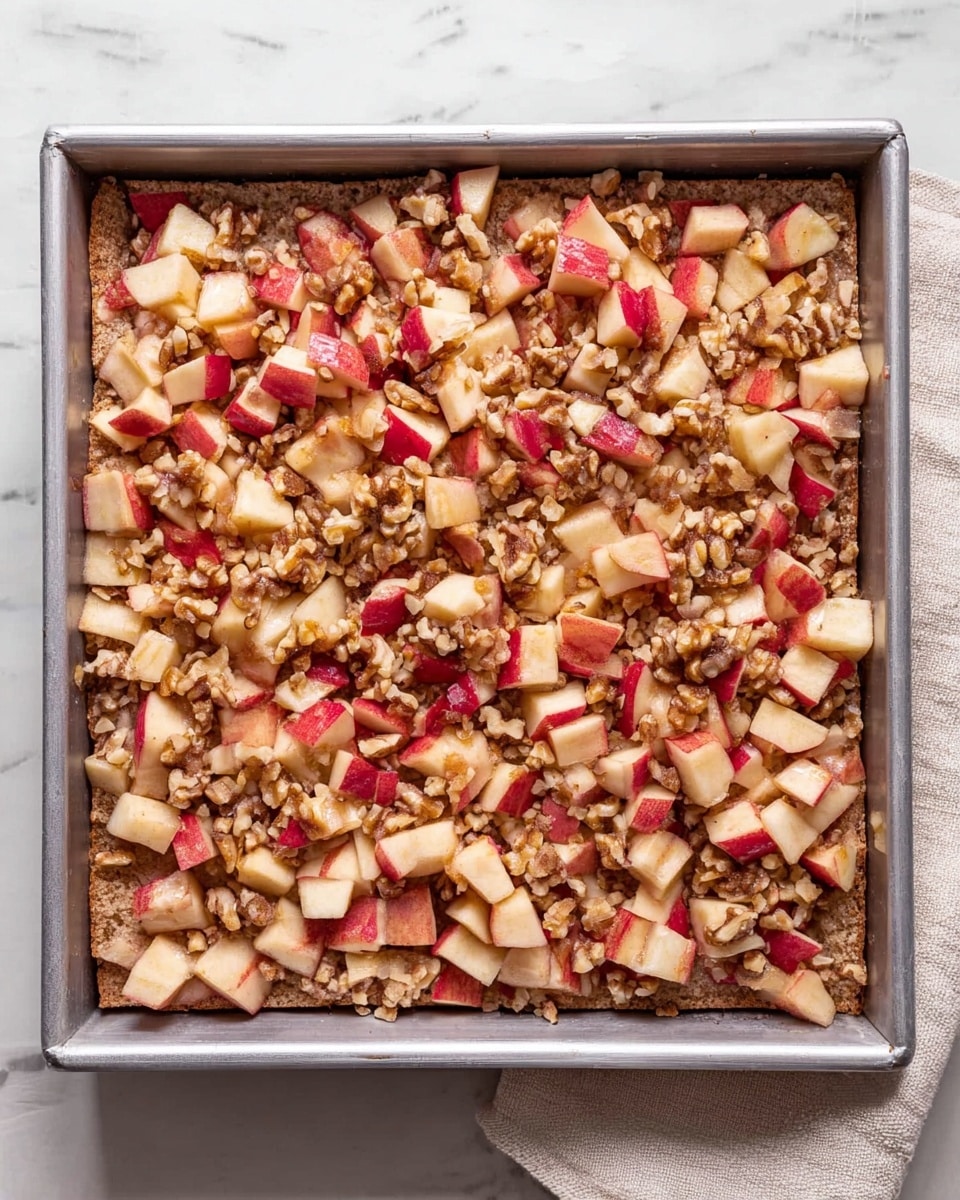 The image shows a square metal baking pan filled with a layered dessert. The bottom layer appears to be a soft, light brown bread or cake base. On top of this base is a thick layer of small, evenly cut apple pieces with red skin and white flesh, mixed with small chunks of walnuts that add a rough texture. The colors are warm and natural, with the reds from the apple skin contrasting with the light browns of the bread and nuts. The baking pan is placed on a white marbled surface, with a light beige cloth partially visible on the right side. photo taken with an iphone --ar 4:5 --v 7