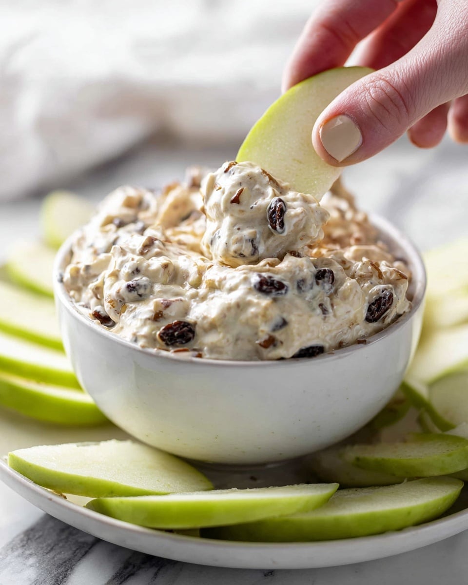 A white bowl filled with a thick creamy dip that has mixed nuts and dark raisins, showing texture and chunks mixed in the dip. A woman’s hand is holding a green apple slice dipped halfway into the creamy mix inside the bowl. The bowl is placed on a white plate, and there are more green apple slices around it, all set on a white marbled surface. The photo has a close-up focus on the dip and the apple slice. photo taken with an iphone --ar 4:5 --v 7