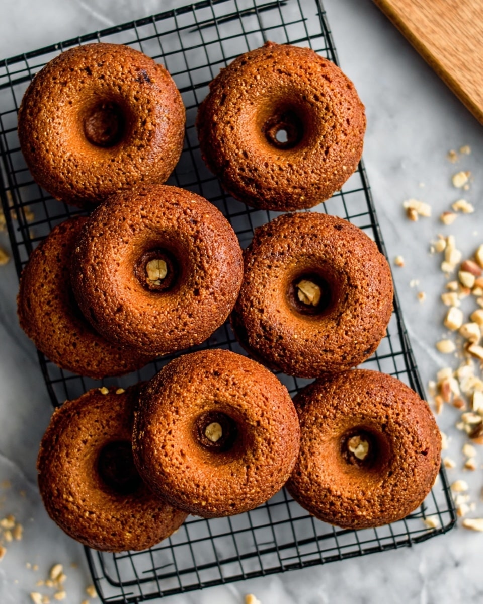 A pile of nine round baked donuts with a medium brown color and a slightly rough texture sits on a black cooling rack. The donuts have a small hole in the center and appear thick with a crunchy outside. The black rack rests on a white marbled surface that has small pieces of nuts scattered near the top right corner. A wooden cutting board is partially visible in the upper right corner. photo taken with an iphone --ar 4:5 --v 7