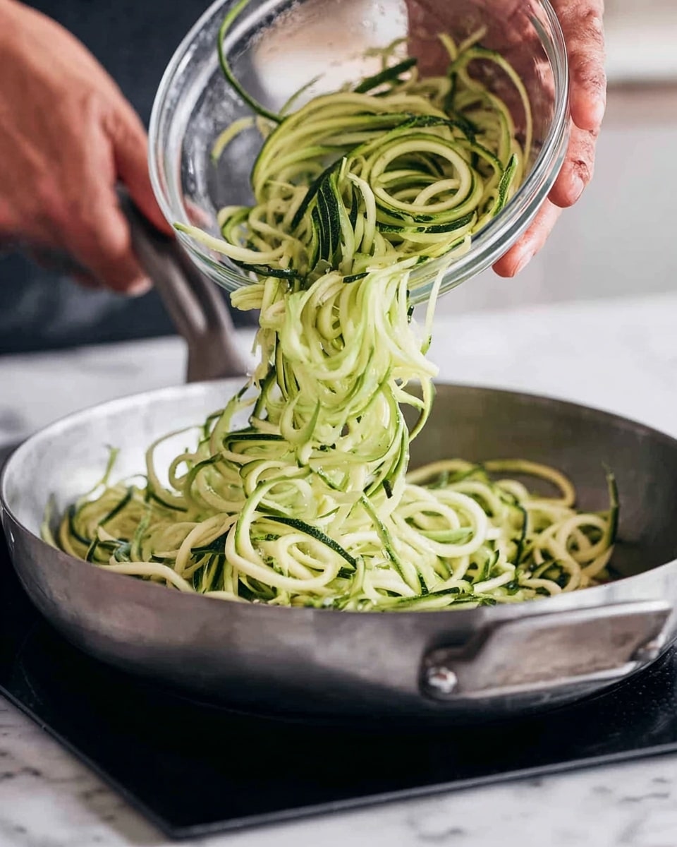 A close-up image showing a person's hands holding a clear glass bowl and pouring green zucchini spiral noodles into a metal frying pan. The zucchini noodles are thin, curly, and fresh-looking with light and dark green shades. The frying pan sits on a black cooking surface, with the background showing a white marbled texture. The person's pale skin and veins are visible on their hands. photo taken with an iphone --ar 4:5 --v 7