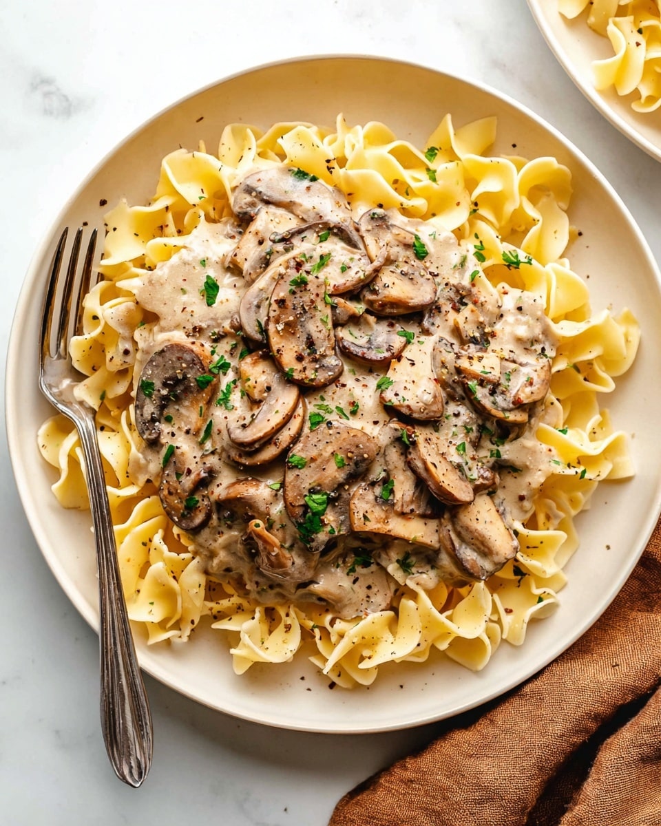 A white round plate holds a serving of wide, ruffled egg noodles that form the bottom layer with a pale yellow color and soft texture. On top, there is a thick layer of creamy mushroom sauce with visible slices of cooked brown mushrooms coated in a light beige sauce, sprinkled with small pieces of fresh green herbs. The dish is seasoned with black pepper flakes scattered over the sauce, and a silver fork rests inside the noodles on the left side of the plate. The background is a white marbled surface with a soft brown cloth partially visible at the bottom right corner. Photo taken with an iphone --ar 4:5 --v 7