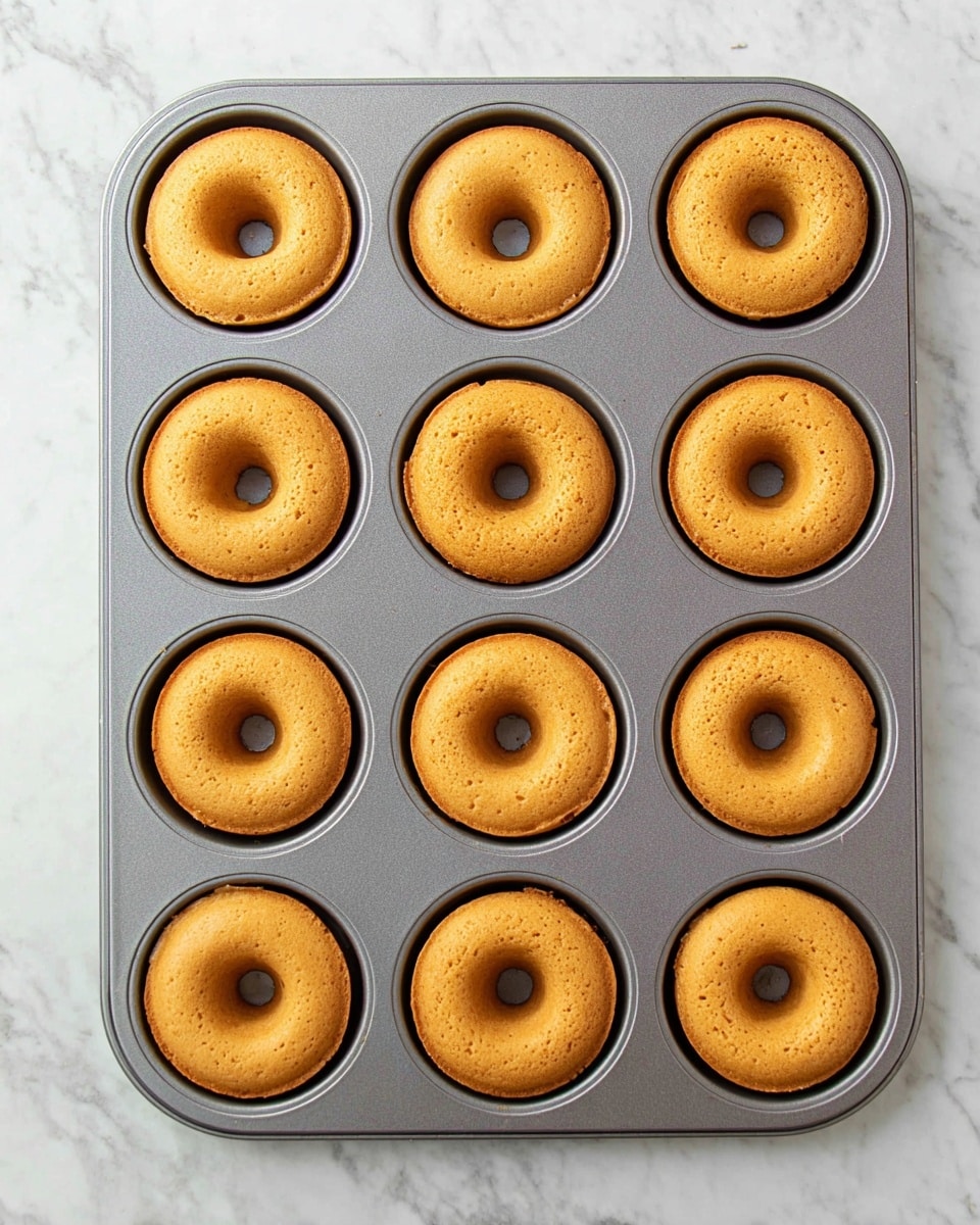 A gray metal donut pan holds twelve freshly baked donuts arranged in three rows and four columns against a white marbled surface. Each donut is golden brown with a smooth, slightly spongy texture and a perfect hole in the center. The donuts are evenly cooked, with a consistent color and a few small air bubbles visible on their surfaces. The pan sits flat and takes up most of the image, giving a clear top view of the donuts inside. photo taken with an iphone --ar 4:5 --v 7