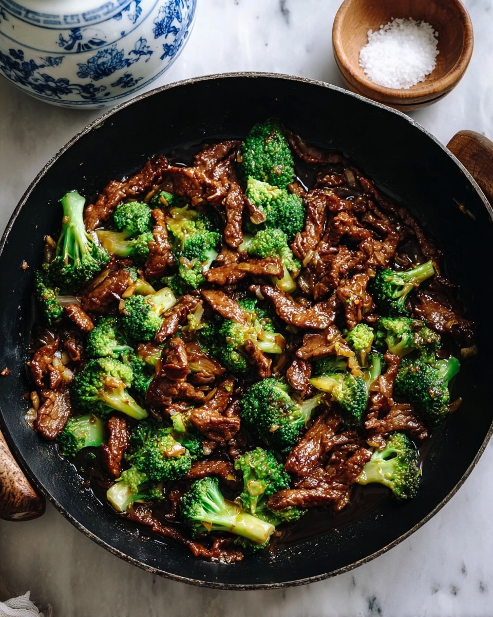 A black pan filled with a stir-fry dish showing two main layers: the bottom layer is dark brown cooked meat strips with a glossy texture from the sauce, and the top layer has bright green broccoli florets scattered evenly across the meat. The meat has a slight caramelized look and some small bits of garlic or ginger mixed in. Around the pan, there is a small wooden bowl with coarse salt and a white ceramic container with blue floral patterns. The background is a white marbled texture. photo taken with an iphone --ar 4:5 --v 7