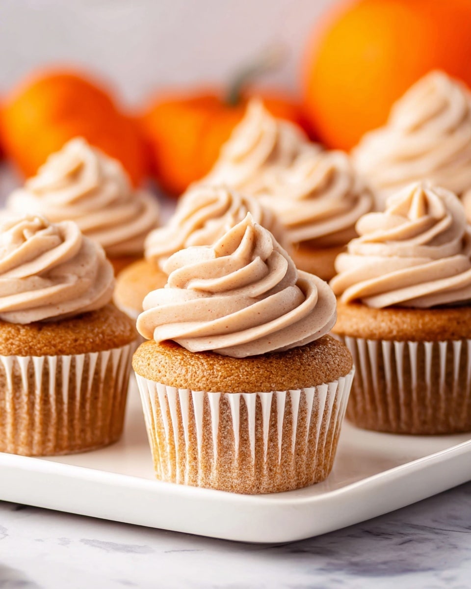 The image shows several golden-brown cupcakes arranged on a white rectangular plate placed on a white marbled surface. Each cupcake has one layer of fluffy, light brown frosting swirled on top in a tall, smooth spiral with soft peaks. The cupcakes have a slightly textured, moist cake base with a ribbed white paper liner around the sides. In the background, there are blurred orange pumpkins adding a warm, autumn feel to the composition. photo taken with an iphone --ar 4:5 --v 7