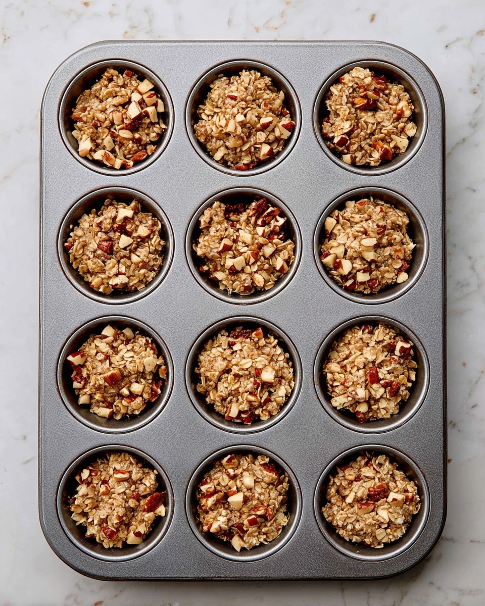 A gray metal muffin tray with twelve cups is filled with unbaked oat and nut mixture. Each cup holds a thick layer of rough-textured oats mixed with small chunks of light brown nuts and pieces of red and tan apple. The oat mixture is uneven and chunky, filling each cup to just below the top edge. The tray is placed on a white marbled surface, and no woman's hand is visible in the image. photo taken with an iphone --ar 4:5 --v 7