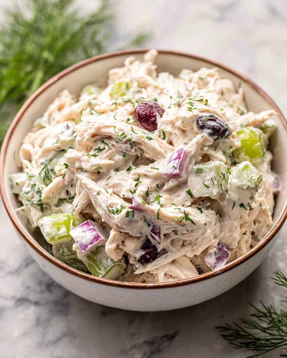 A close-up view of a bowl filled with creamy chicken salad showing a mix of shredded white chicken pieces, small cubes of purple-red onion, slices of green celery, and some darker bits that look like olives or grapes, all coated in a thick white sauce with small green herb sprinkles on top. The bowl is white with a thin brown rim, sitting on a white marbled surface, with some green dill sprigs blurred in the background. The texture looks creamy and chunky with visible fresh ingredients mixed evenly. Photo taken with an iphone --ar 4:5 --v 7