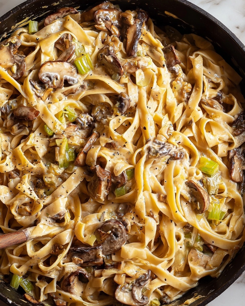 A close-up view of creamy pasta with mushrooms in a black pan. The pasta is long and flat with a pale yellow color, mixed with tender, dark brown mushroom slices and small bits of light green celery. The creamy sauce coats the pasta and vegetables evenly, making the dish look smooth and rich. The textures are soft with some slight gloss from the sauce, and a few black pepper specks add small dark spots. The pan’s black edges frame the warm-toned dish, sitting on a white marbled surface. photo taken with an iphone --ar 4:5 --v 7