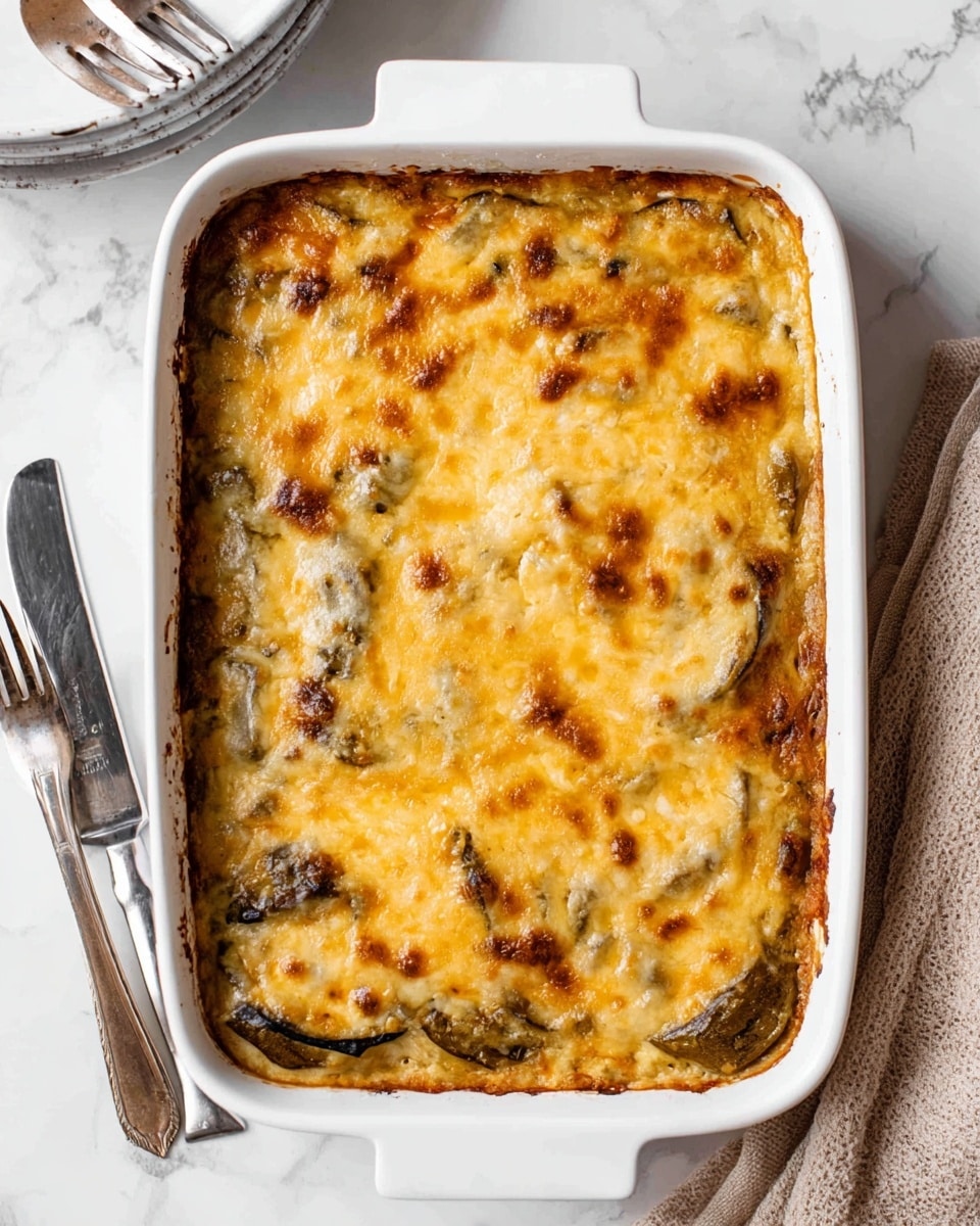 A baked dish in a white rectangular baking pan with a golden brown, melted cheese layer on top, showing spots of light and darker yellow cheese with some browned edges; under the cheese, slices of cooked vegetables, likely eggplant, create uneven greenish-brown patches; the dish appears creamy and soft with a bubbly, slightly crispy top layer. The baking pan sits on a white marbled surface with a fork and knife on the left and a beige cloth napkin partially visible on the right. Photo taken with an iphone --ar 4:5 --v 7
