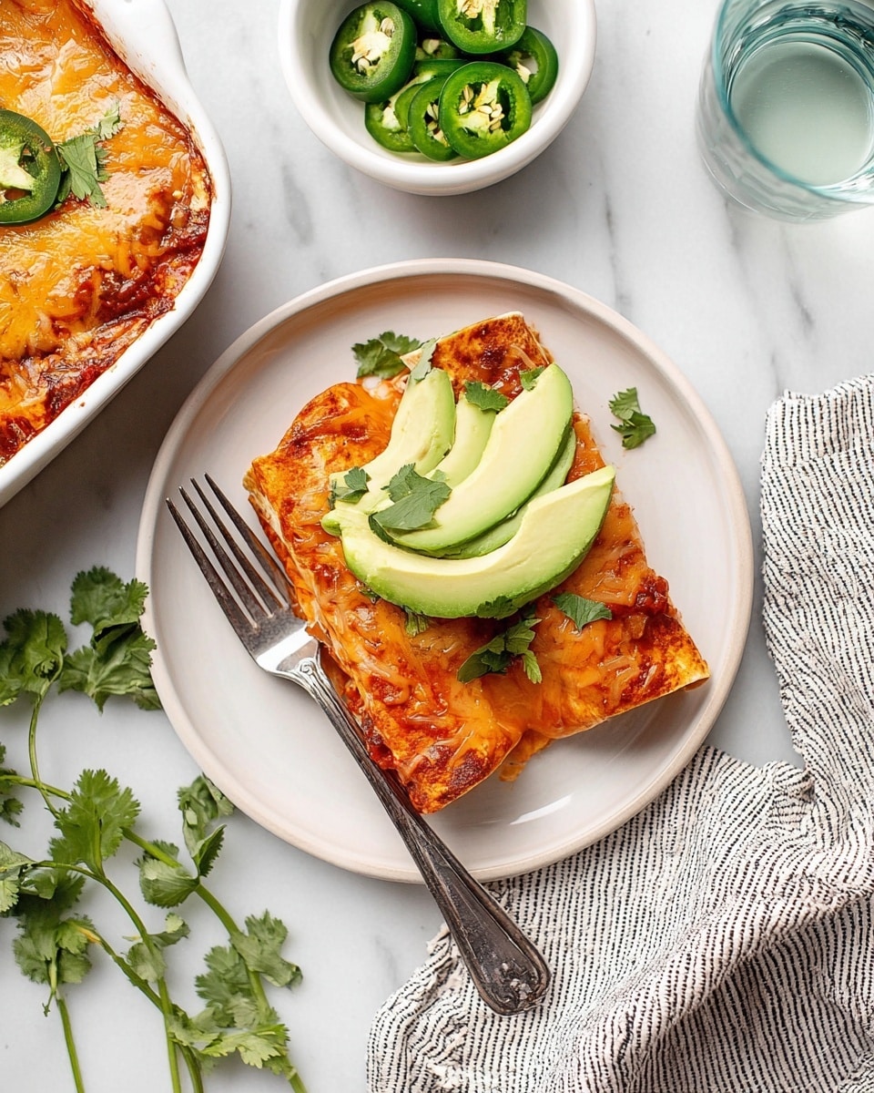 A white plate on a white marbled surface holds two stacked enchiladas covered in melted orange-red cheese and sauce, topped with four thin slices of bright green avocado and a sprig of fresh cilantro. A silver fork rests under the enchiladas with its handle pointing outward. To the left, part of a white dish of enchiladas topped with cheese, avocado slices, and green jalapeño slices is visible. There is also a small white bowl filled with sliced green jalapeños above the plate, fresh cilantro leaves scattered around, and a clear glass of water with a light blue tint on the right. A white and black striped cloth is draped near the plate. Photo taken with an iphone --ar 4:5 --v 7