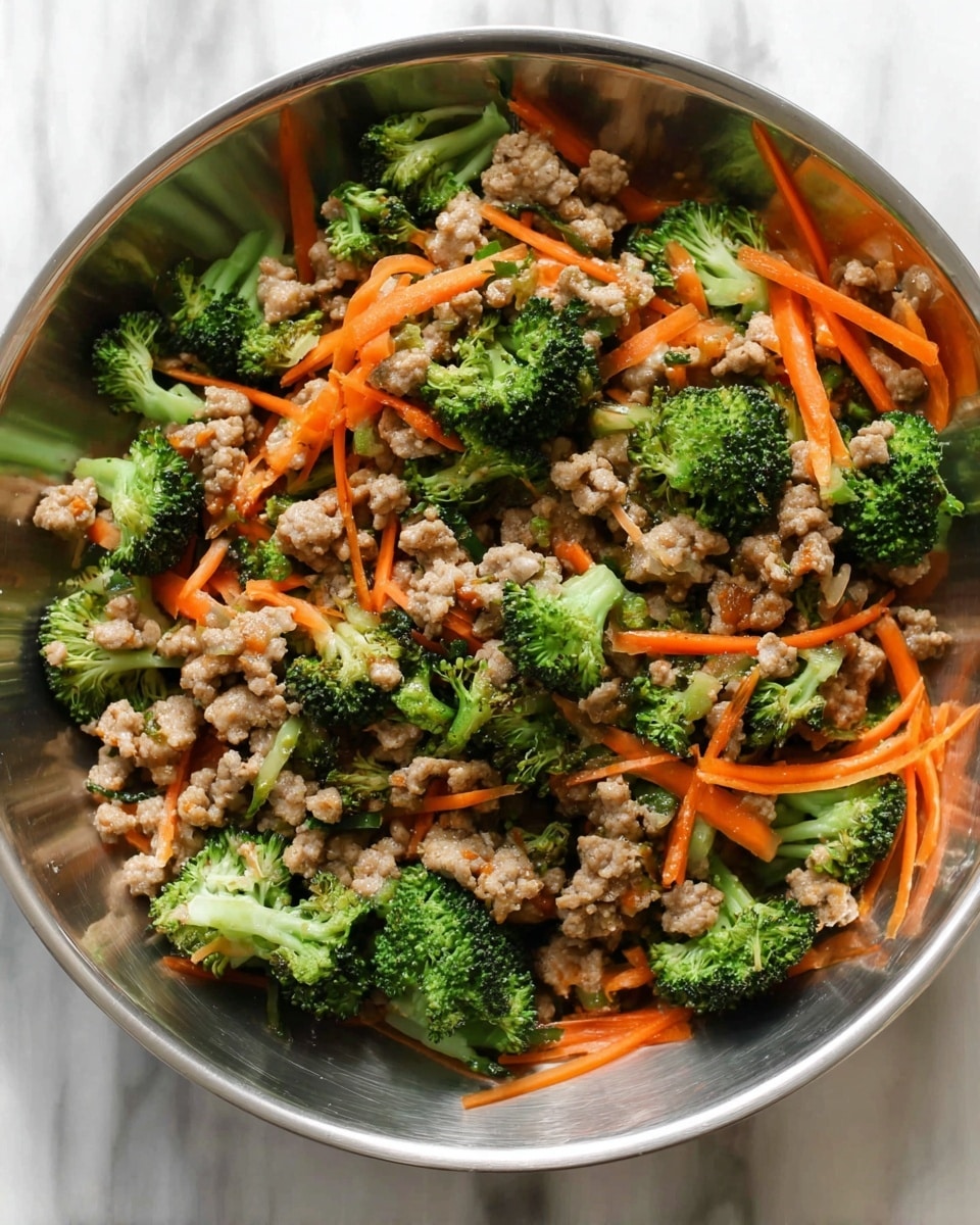 The dish is shown in a large silver bowl filled with a mix of cooked ground meat, bright green broccoli florets, and thin bright orange carrot strips. The ground meat is light brown with a slightly crumbly texture, spread evenly throughout. The broccoli pieces are fresh and chunky, placed on top and mixed within the meat, while the carrot strips are scattered thin and long across the whole dish. There are small drops of sauce that add a slight shine to the ingredients, making the colors look richer. The background is a white marbled texture. photo taken with an iphone --ar 4:5 --v 7