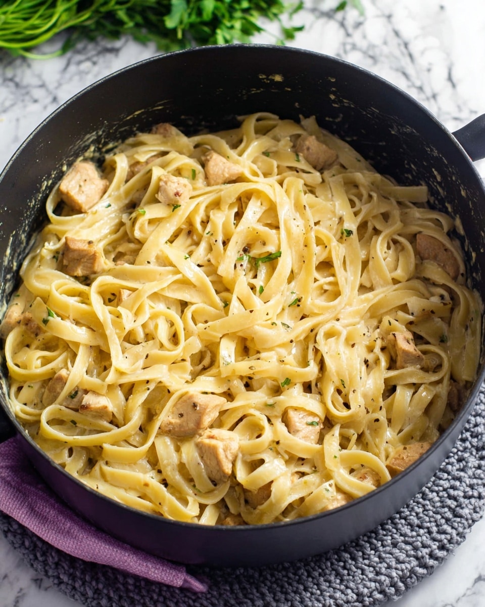 The image shows a black pot filled with creamy fettuccine pasta mixed with small pieces of light brown chicken. The pasta is long and flat with a smooth texture, coated in a pale yellow sauce with tiny black pepper flakes. The chicken pieces are scattered evenly through the pasta, appearing tender and juicy. The pot sits on a knitted grey mat, and the background features a white marbled surface with green herbs slightly blurred in the distance. Photo taken with an iphone --ar 4:5 --v 7