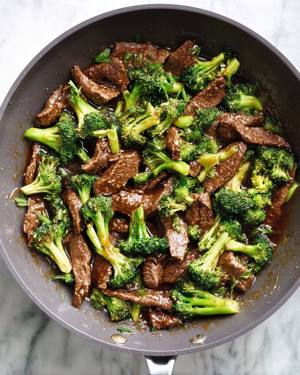 A grey pan filled with cooked beef slices and bright green broccoli pieces, all covered in shiny brown sauce. The beef pieces are thin and tender looking, mixed evenly with fresh broccoli florets that have light green stems. The sauce has a smooth texture, coating the meat and vegetables lightly, giving a glossy and moist appearance. The pan sits on a white marbled surface. photo taken with an iphone --ar 4:5 --v 7