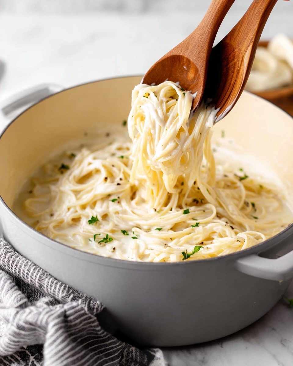 A pot filled with creamy white spaghetti noodles coated in a smooth, thick white sauce, with small green herb pieces scattered on top. Two wooden spoons are lifting a bunch of noodles from the middle, showing the soft, creamy texture of the pasta. The pot is placed on a white marbled surface, with a gray-striped cloth near its edge. The background is softly blurred with white tones, highlighting the pasta as the main focus. Photo taken with an iphone --ar 4:5 --v 7