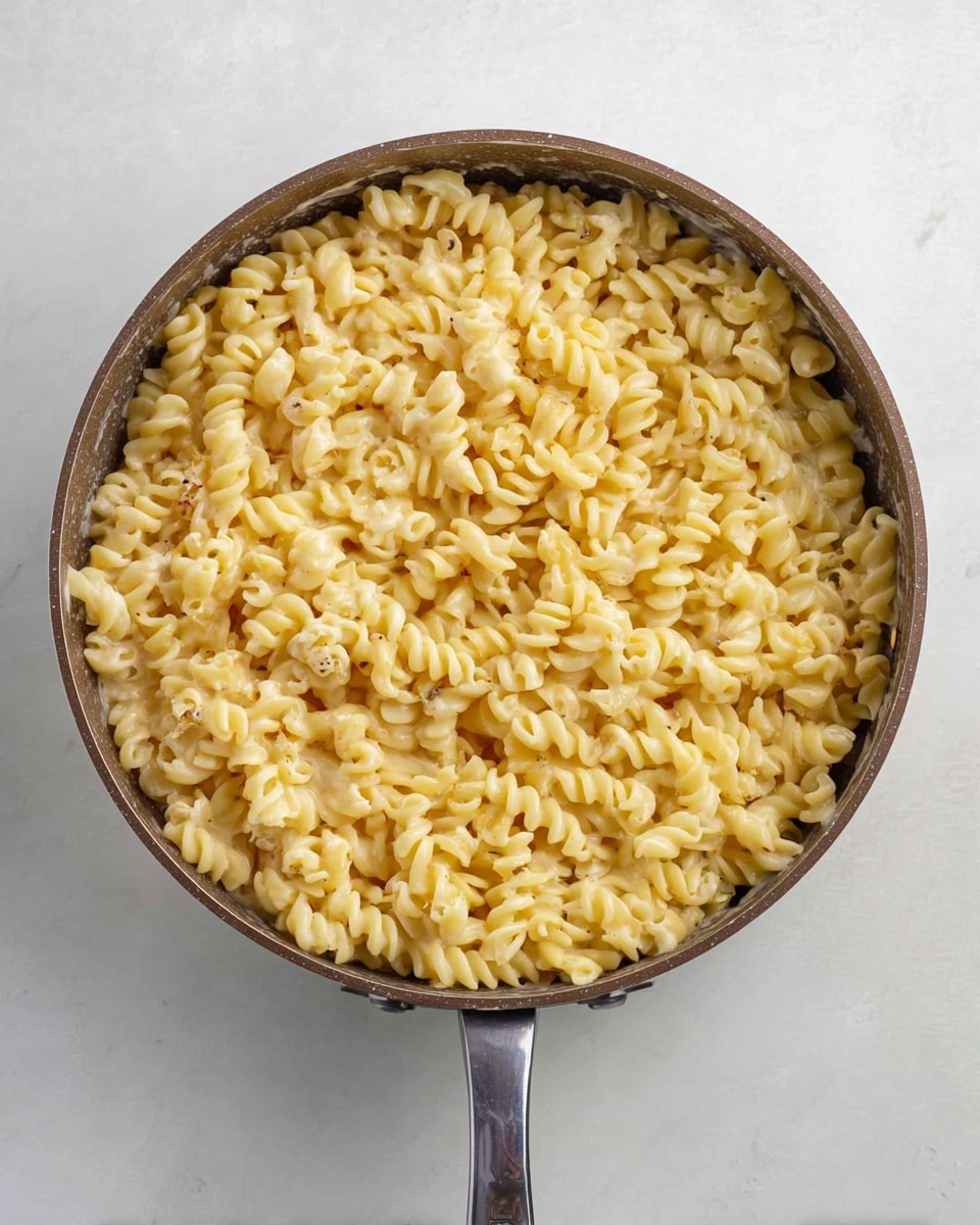 This image shows a deep light brown pan filled with cooked curly pasta on a white marbled surface. The pasta is light yellow and appears creamy, with a few small pieces of light cream-colored sauce or cheese mixed in. The pasta fills the pan evenly from edge to edge, and the pan has a handle extending downward, centered in the frame. The texture looks smooth and soft, with a slight shine from the sauce. Photo taken with an iphone --ar 4:5 --v 7