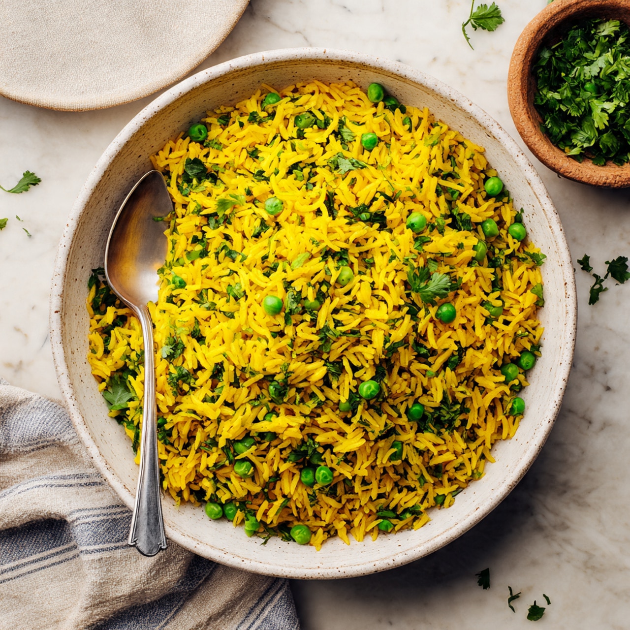 A large white speckled bowl filled with yellow rice mixed with bright green peas and fresh chopped green herbs, mostly cilantro. The rice looks soft and fluffy with small spices visible throughout. A silver spoon rests inside the bowl on the left side. The bowl sits on a white marbled surface with some scattered small green herb leaves. A striped cloth napkin is partially visible at the top left corner and a small brown bowl filled with green cilantro leaves is at the top right corner of the image, photo taken with an iphone --ar 4:5 --v 7