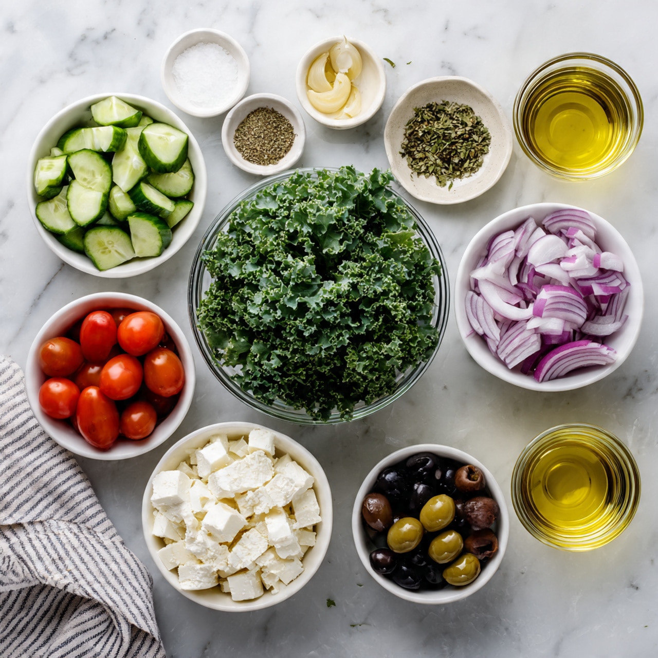 The image shows several small white bowls arranged on a white marbled surface, each holding a different ingredient. In the center, a clear glass bowl contains bright green curly kale leaves. Surrounding it are white bowls filled with chopped green cucumbers, halved red cherry tomatoes, chopped purple onions, whole black olives, and crumbled white cheese. Smaller white bowls hold minced garlic, dried herbs, salt, and black pepper. Two small glasses with golden and light yellow liquids are also present. A striped cloth napkin rests on the side. Photo taken with an iphone --ar 4:5 --v 7