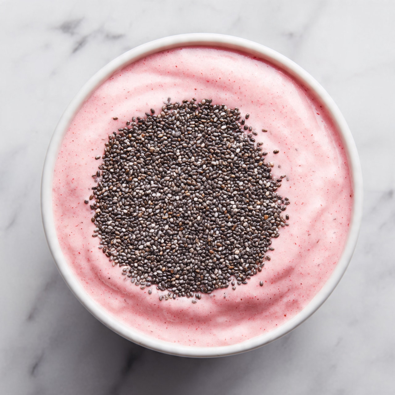 A clear white bowl holds a pink creamy smoothie layer spread evenly inside, topped with a dense, circular layer of tiny black chia seeds that cover most of the surface. The bowl is set on a white marbled surface, showing a smooth texture with the chia seeds creating a rough, grainy contrast against the soft pink base, photo taken with an iphone --ar 4:5 --v 7