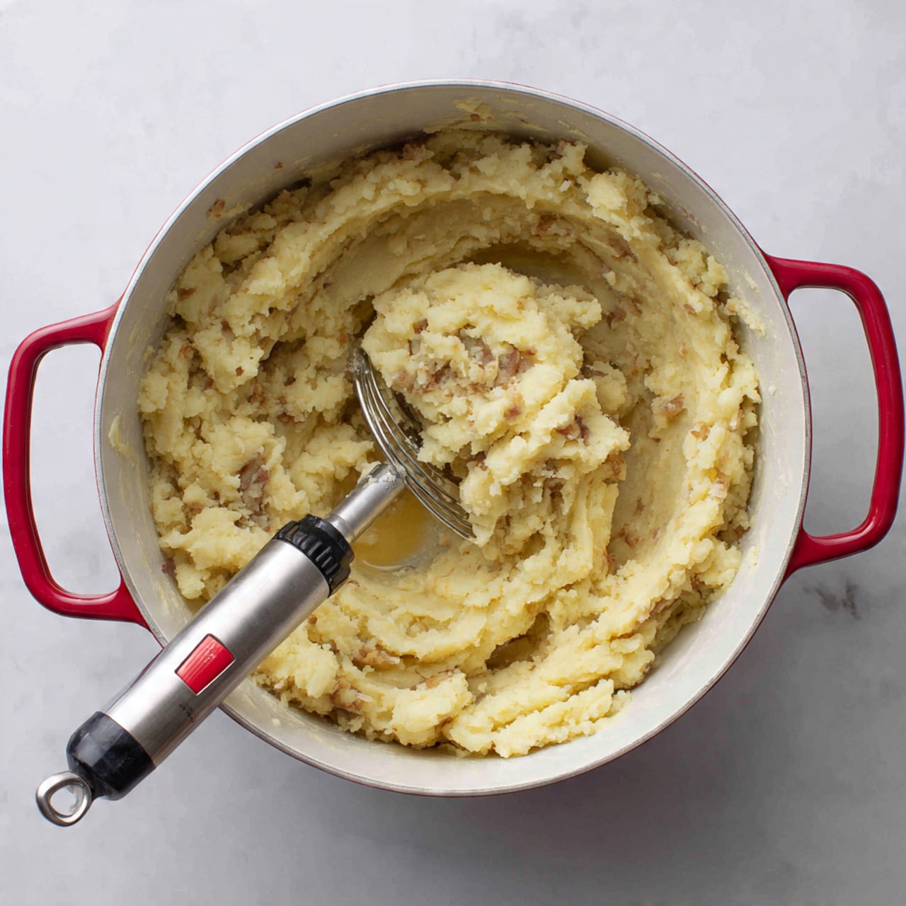 Inside a white pot with red handles, there is a mixture of mashed potatoes being mashed with a silver masher that has a black handle with a red spot. The mashed potatoes show chunks of light yellow potato with bits of brown potato skin mixed inside. A small amount of white liquid is visible among the creamy texture. The pot sits on a white marbled surface. photo taken with an iphone --ar 4:5 --v 7