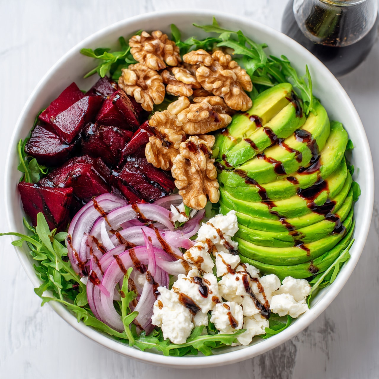 A white bowl filled with a fresh salad on a white marbled surface. The bottom layer is dark green leafy arugula. On top of that, there are deep red beet cubes placed mainly on one side. Next to the beets are thin slices of light purple-red onion rings. On the other side, there are bright green avocado slices. Scattered on the salad are small white crumbles of cheese and crunchy brown walnut pieces. The salad is drizzled with a dark dressing, giving it a shiny look. Photo taken with an iphone --ar 4:5 --v 7