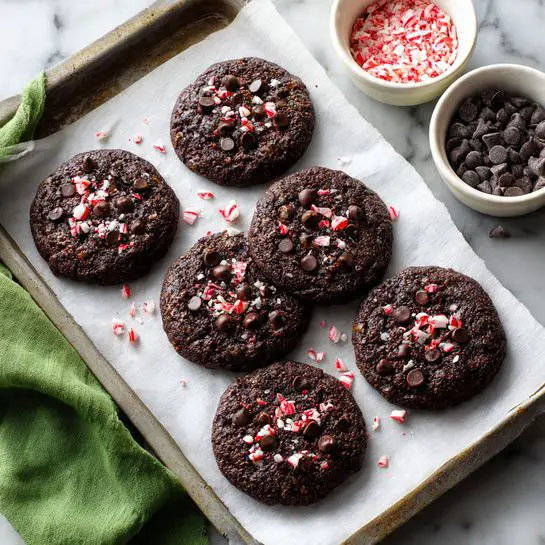 Six dark brown chocolate cookies with a rough texture are placed on a white baking paper on a baking tray. Each cookie is topped with red and white crushed peppermint pieces and dark chocolate chips, scattered unevenly on the surface. The cookies have a slightly uneven round shape, and small bits of peppermint are also scattered around them on the paper. Around the tray, there are parts of bowls filled with dark chocolate chips and crushed peppermint pieces visible, set on a white marbled surface, with a green cloth partly in view on the left side. photo taken with an iphone --ar 4:5 --v 7