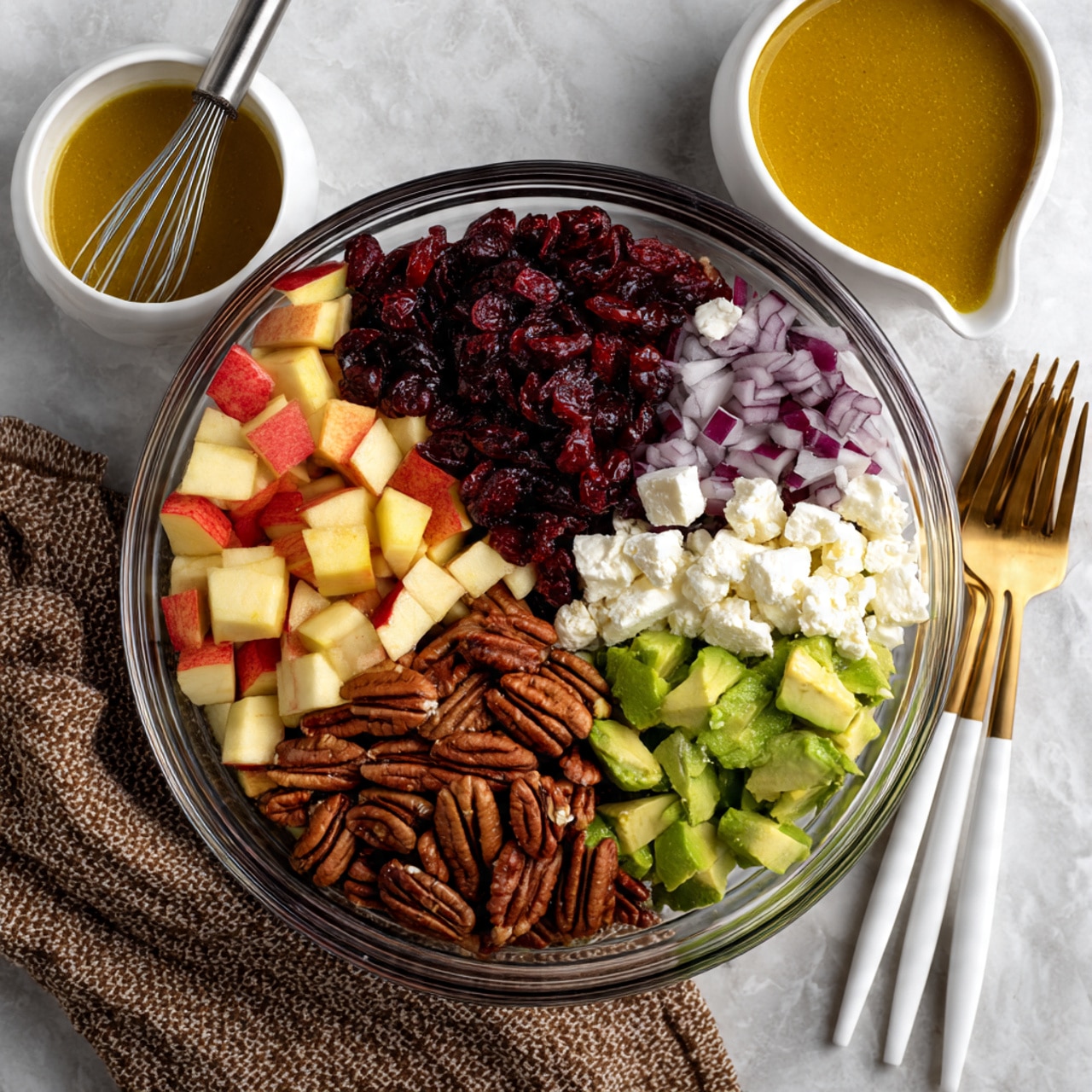 A clear glass bowl sits on a white marble textured surface and holds seven neat sections of ingredients. Starting from the top left and moving clockwise, there are reddish-yellow chopped apple pieces, dark red dried cranberries, finely chopped purple onion, crumbled white feta cheese, green diced avocado, brown pecans, and light beige chickpeas. To the left of the bowl, there is a white bowl with a mustard-yellow dressing and a small whisk inside. To the right, two golden salad forks with white and gold handles rest partly on the marble surface and partly on the bowl edge. The scene is well-lit and clear. photo taken with an iphone --ar 4:5 --v 7