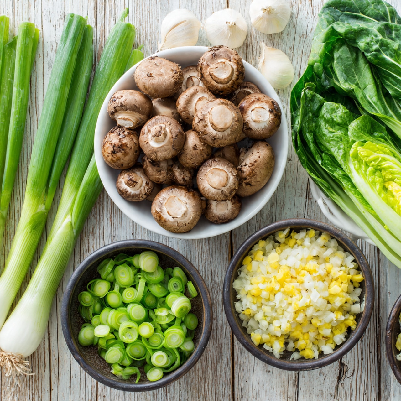 The image shows fresh ingredients arranged on a light wooden surface replaced with a white marbled texture. There are five main items: on the left side, three whole green onions in bright green color lie flat. Next to them, a white bowl filled with sliced brown mushrooms, showing a mix of light and dark brown caps and white stems, takes a center spot. Above and to the right, another white bowl holds fresh, whole green leafy bok choy, with thick pale green stems and dark green leaves. Below it, a smaller dark bowl contains sliced green onions, showing a range of light to dark green pieces. Finally, the last dark bowl at the bottom right is filled with finely chopped ingredients — mostly pale yellow and white, likely garlic and shallots. The layout is clean and organized, the colors mainly various greens, browns, and whites. photo taken with an iphone --ar 4:5 --v 7