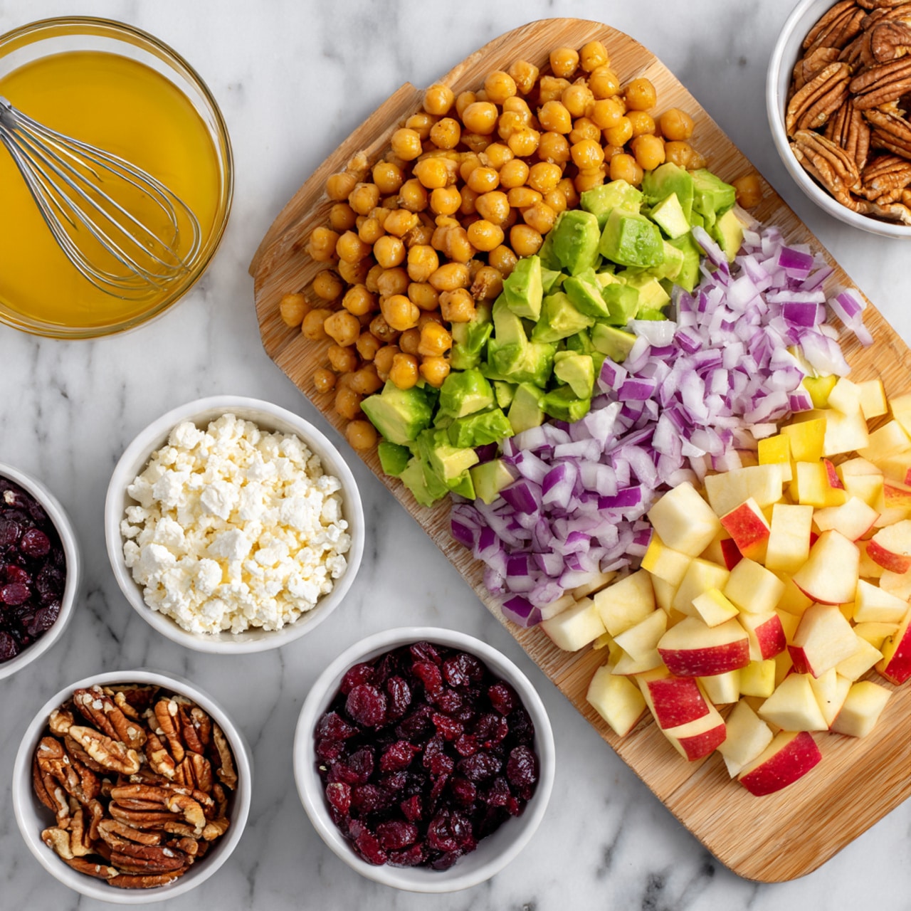 The image shows several small bowls and a white cutting board on a white marbled surface. The cutting board has three layers: diced red onion with purple and white colors at the top, bright green diced avocado in the middle, and light yellow and red diced apple pieces at the bottom. Next to it are five small white bowls, each with a different ingredient: golden brown chickpeas in one large bowl, crumbled white cheese in a small bowl, dark red dried cranberries in another small bowl, and chopped pecans in a small bowl. At the top is a bowl with a golden yellow liquid dressing and a small whisk inside it. photo taken with an iphone --ar 4:5 --v 7
