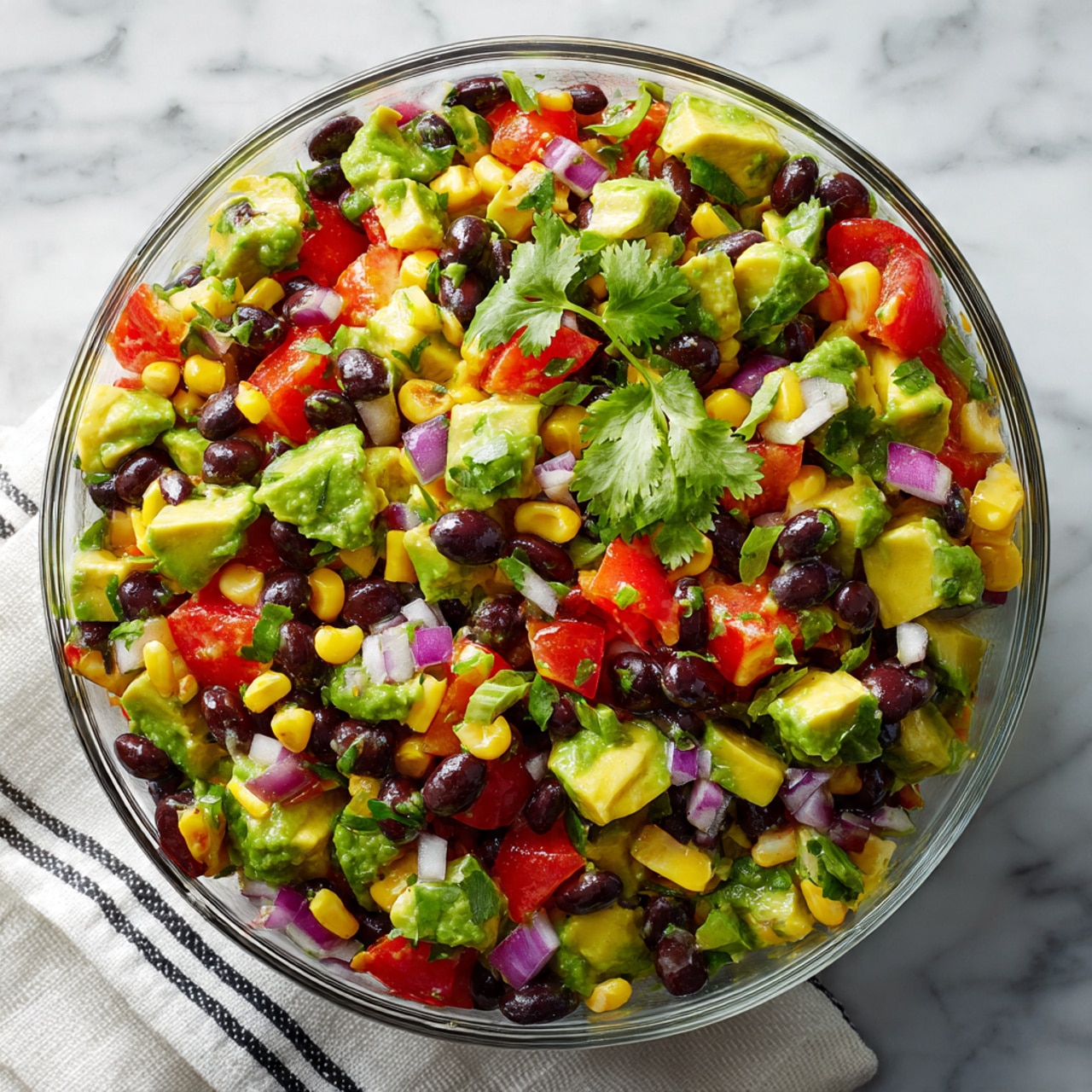 The image shows a clear glass bowl filled with a colorful mixed salad. The salad has several layers: bright green chunky avocado pieces, black beans scattered throughout, yellow corn kernels, diced red tomatoes, small bits of purple onion, and a few green cilantro leaves on top. The bowl sits on a white marbled surface with a white cloth that has black stripes underneath. The overall look is fresh and vibrant with a mix of green, black, yellow, red, and purple colors. photo taken with an iphone --ar 4:5 --v 7