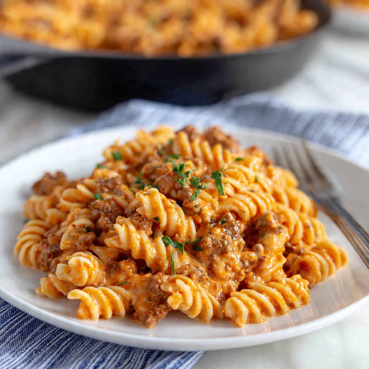 The image shows a white plate with a single pile of spiral pasta coated in a thick, creamy, orange-colored sauce mixed with ground meat. The pasta has a slightly shiny texture and is sprinkled with small green herb pieces on top. Next to the plate on a blue and white striped cloth lies a silver fork with a long black handle. In the background, a black skillet filled with more of the pasta and sauce sits on a white marbled surface. Photo taken with an iphone --ar 4:5 --v 7