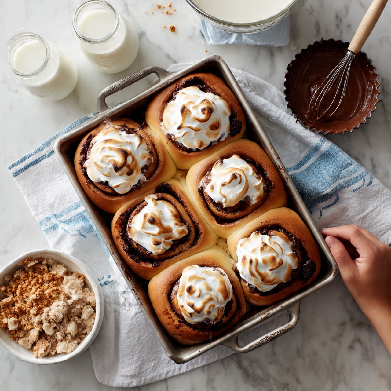 In a rectangular metal baking pan, there are six warm rolls arranged in two rows of three, each roll showing distinct layers of light brown dough spiraled with dark chocolate. On top of each roll, there is a thick, toasted layer of white meringue with golden brown spots and swirls. The rolls look soft and slightly glossy, with melted chocolate peeking through the dough layers. The pan sits on a white marbled surface next to a white cloth with blue lines, a small round tart pan filled with melted dark chocolate, a metal bowl holding broken golden brown crumbs, and a white lacy plate partially visible at the bottom. In the background, a whisk covered in white meringue rests in a white bowl, with two glasses of milk behind it. Photo taken with an iphone --ar 4:5 --v 7