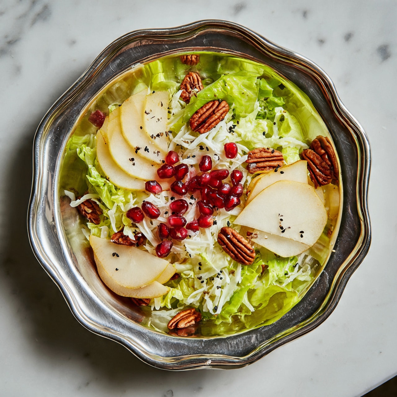 A salad is served in a shiny silver dish with a wavy edge, placed on a white marbled surface. The bottom layer is made of fresh green lettuce leaves, topped with a light layer of shredded white cheese. On top, there are bright red pomegranate seeds scattered across, and pieces of brown pecans are spread evenly. Thin slices of light yellow pear are arranged in small groups around the salad, sprinkled with tiny black seeds. The colors are bright and fresh, making the salad look healthy and tasty. Photo taken with an iphone --ar 4:5 --v 7