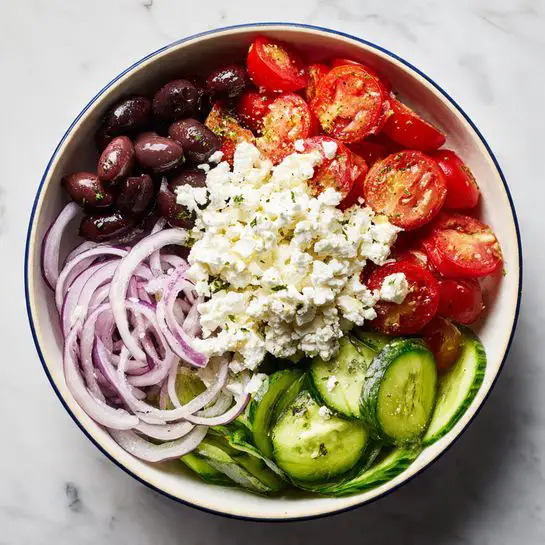 A bowl contains a fresh salad with four distinct layers divided into sections: dark purple sliced olives on the top left, bright red halved cherry tomatoes on the top right, thin light purple onion rings overlapping the bottom right, and thinly sliced green cucumber rounds on the bottom left. In the center, a generous pile of white crumbly feta cheese rests on top, slightly spilling onto the other ingredients. The bowl is white with a thin blue rim and sits on a white marbled surface. photo taken with an iphone --ar 4:5 --v 7