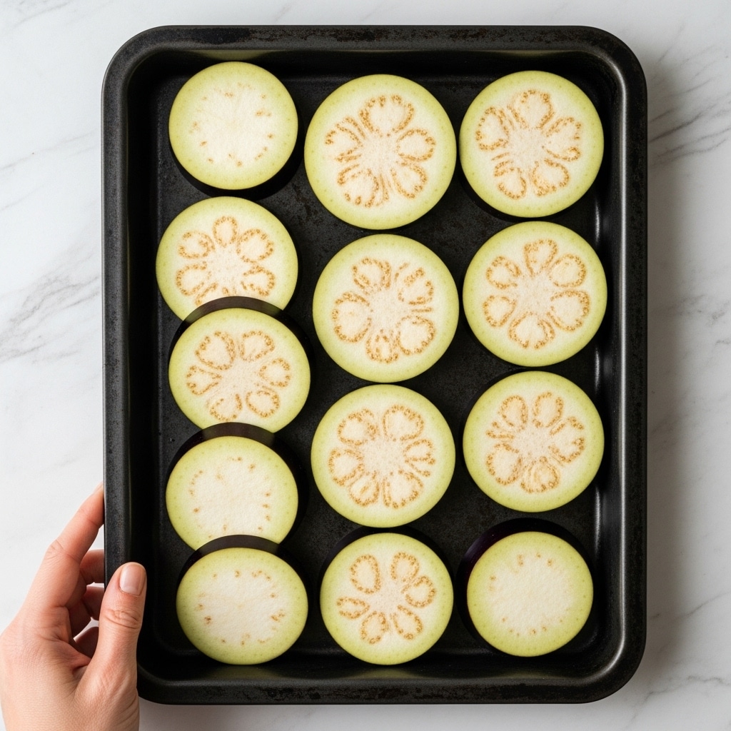A black baking tray holds 13 slices of eggplant arranged in three rows, with the middle row slightly overlapping. The slices are pale white with a soft purple edge and have light brown seeds scattered inside. A woman's hand is gently touching the bottom left corner of the tray. The tray is placed on a white marbled surface. photo taken with an iphone --ar 4:5 --v 7