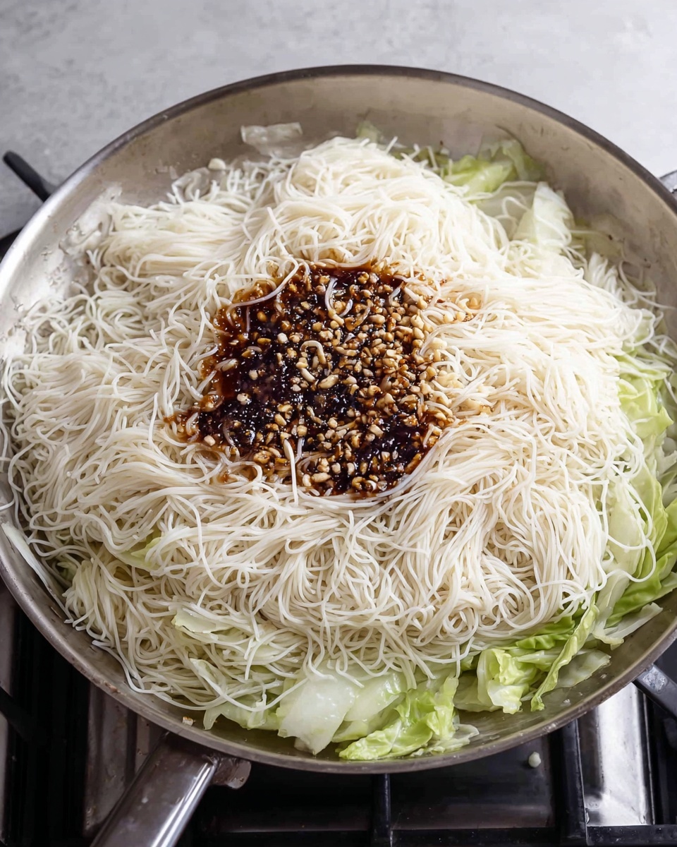 Panda Express Chow Mein Recipe 4 A large silver frying pan filled with a layer of thin, white noodles piled high in the center. On top of the noodles, there is a small pool of dark brown sauce mixed with finely chopped garlic or nuts, creating a textured contrast against the smooth noodles. Underneath the noodles, on one side, some light green cabbage leaves peek out, adding a touch of color. The pan is placed on a stove with a grey surface partially visible. photo taken with an iphone --ar 4:5 --v 7