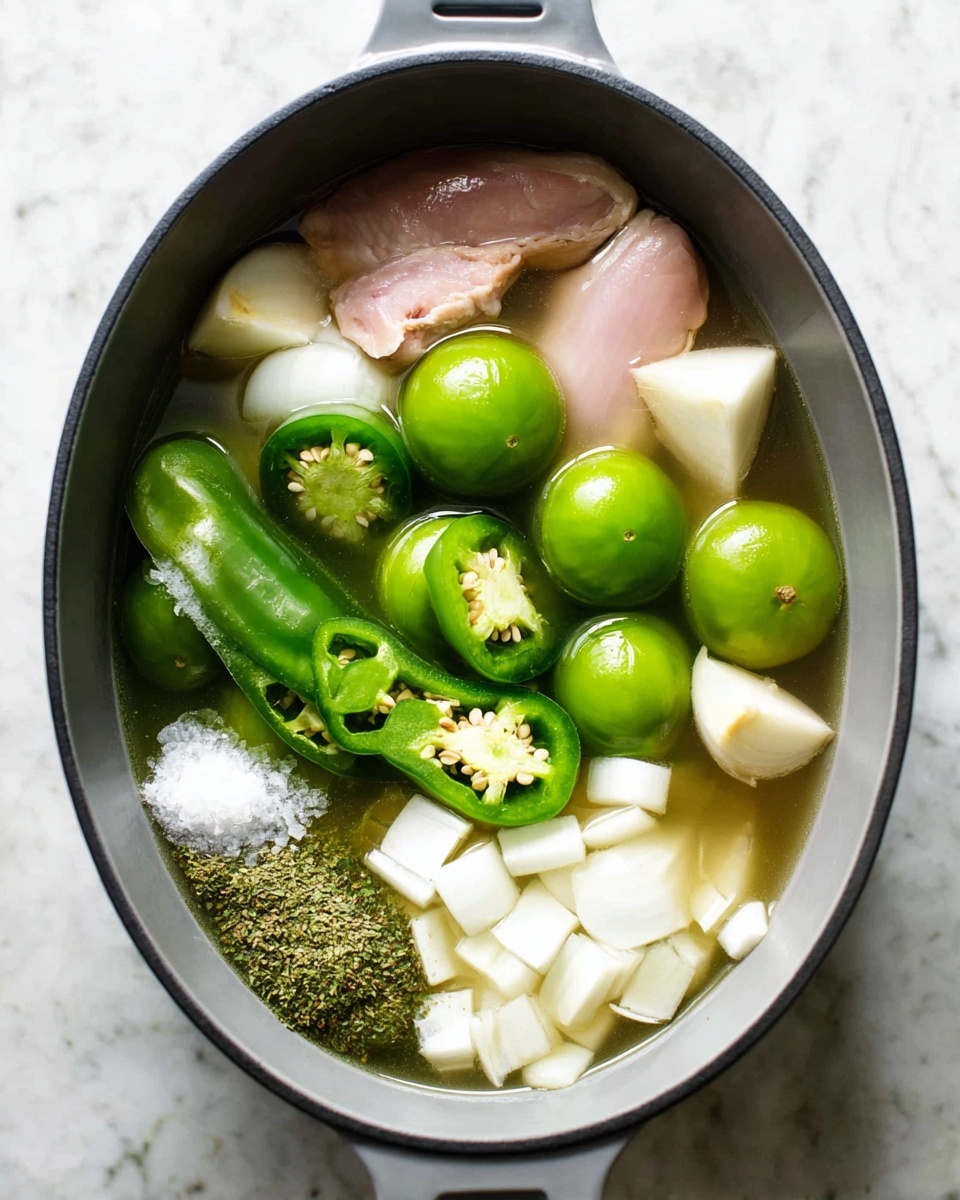 A large oval gray pot filled with a clear broth holding several whole bright green tomatillos floating on top. There are chunks of white onion scattered throughout, along with three large green jalapeño halves with visible seeds, and two peeled garlic cloves in the center. Two raw pale pink chicken pieces sit partially submerged in the broth near the bottom left. A small pile of dried green herbs and white salt rests on chopped onion pieces near the bottom right. The pot is set on a white marbled surface. Photo taken with an iphone --ar 4:5 --v 7