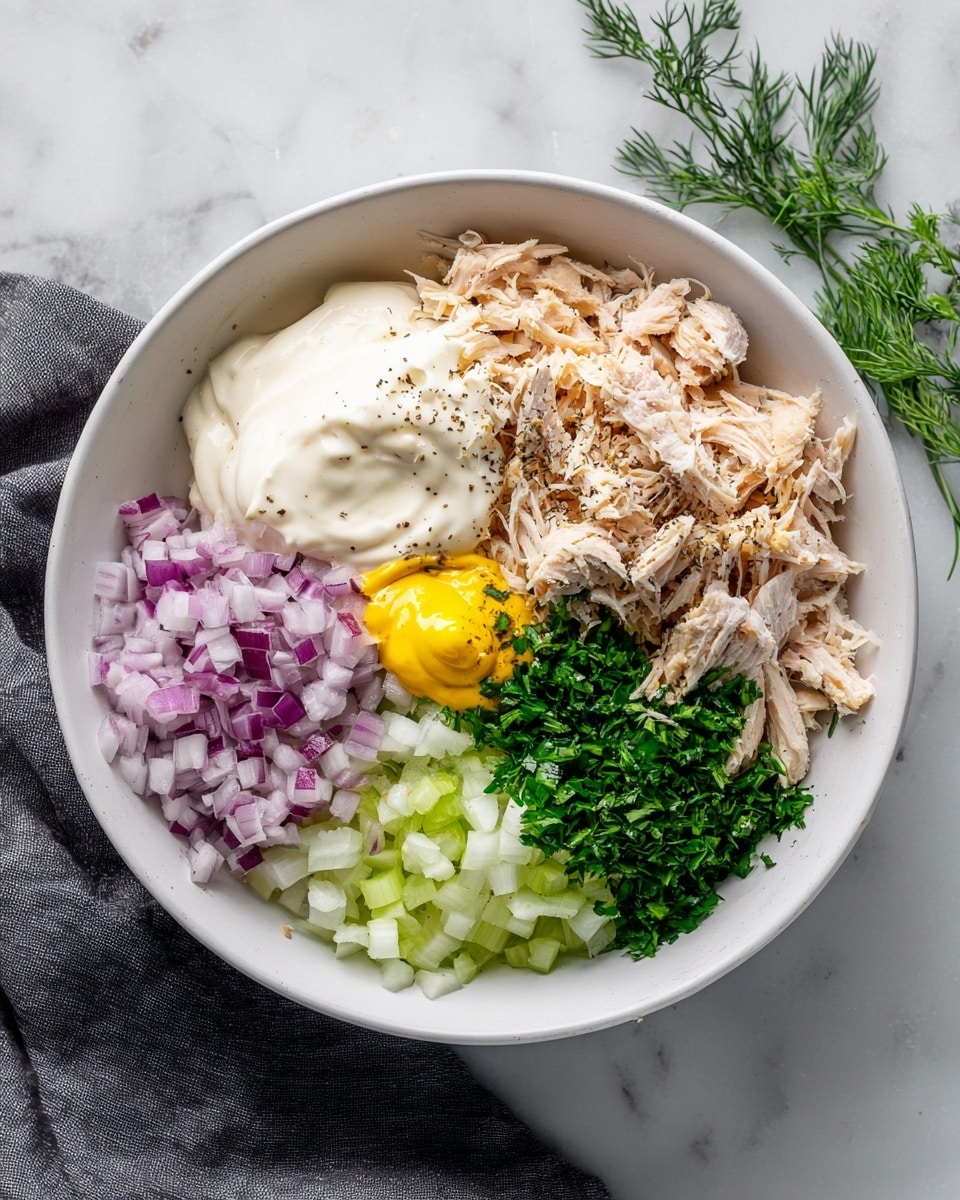 A white bowl filled with several layered ingredients on a white marbled background. Starting from the top right, there is a layer of shredded light brown cooked chicken with a sprinkle of black pepper on part of it. Below the chicken is a small dollop of bright yellow mustard next to a large creamy white mayonnaise portion. To the left of the mayonnaise is a section of finely chopped purple onions, and above that is a layer of light green chopped celery. In the center of the bowl, there is a small pile of finely chopped fresh green herbs. The bowl is placed near some green herb sprigs and a dark gray cloth on the white marbled surface. Photo taken with an iphone --ar 4:5 --v 7