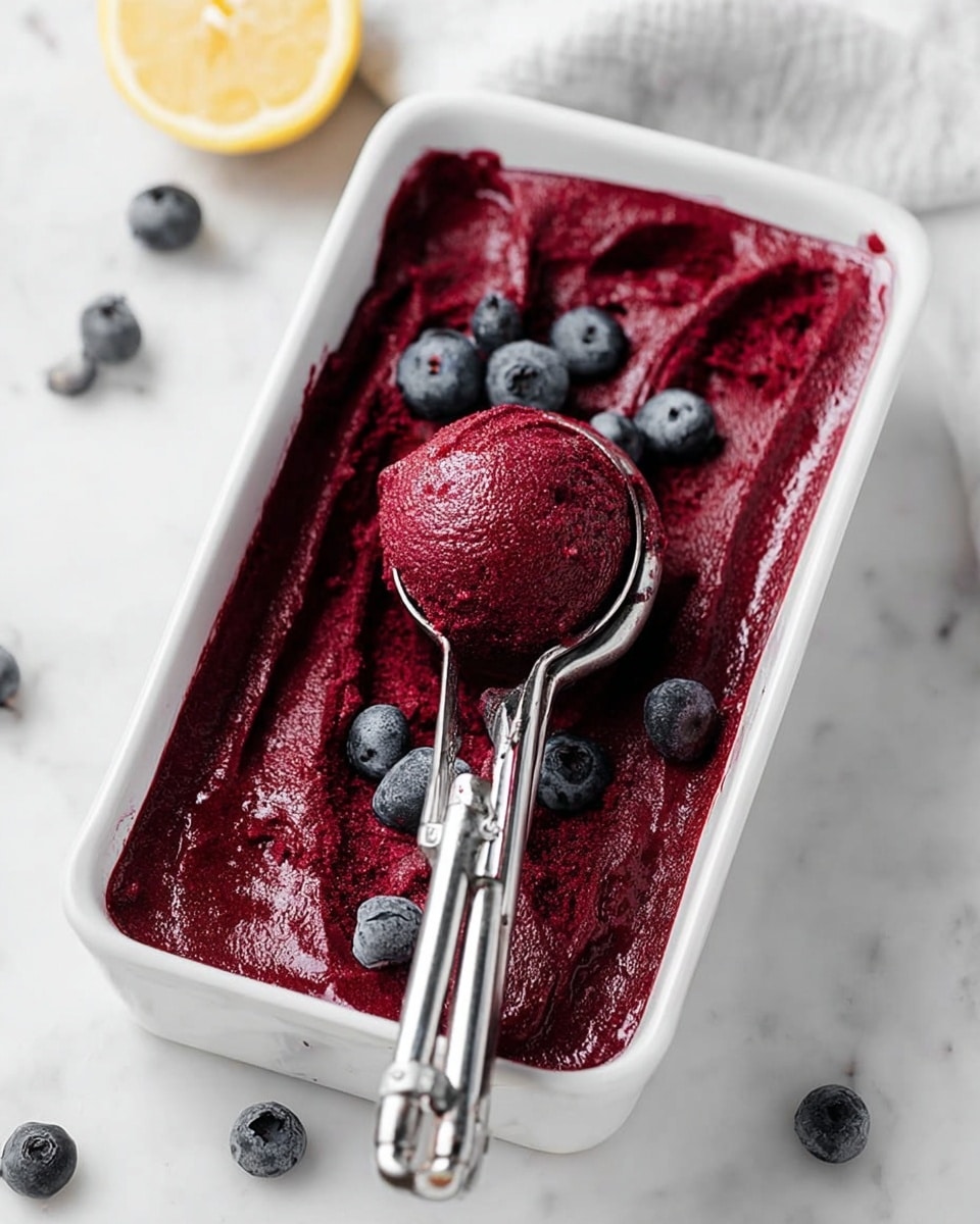 A white rectangular dish filled with a deep red-purple smooth ice cream or sorbet, with a shiny texture and slight ripples on the surface. There is a metal ice cream scoop resting on the dish, holding a round scoop of the sorbet. Scattered on top around the scoop are several fresh blueberries with a soft blue-gray color. The dish sits on a white marbled surface with a half lemon visible on the top left corner and a few loose blueberries scattered around. Photo taken with an iphone --ar 4:5 --v 7