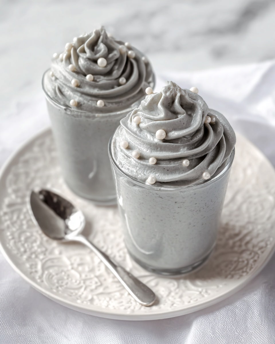 Two clear glasses filled with three layers of smooth, light gray whipped mousse, each topped with a swirl of the same gray mousse decorated with small round white pearl-like sprinkles. The glasses are placed side by side on a white plate with an embossed pattern, sitting on a white marbled surface. A silver spoon lies beside the back glass, resting on the plate. The light is soft, highlighting the creamy texture and subtle shadows of the mousse. Photo taken with an iphone --ar 4:5 --v 7