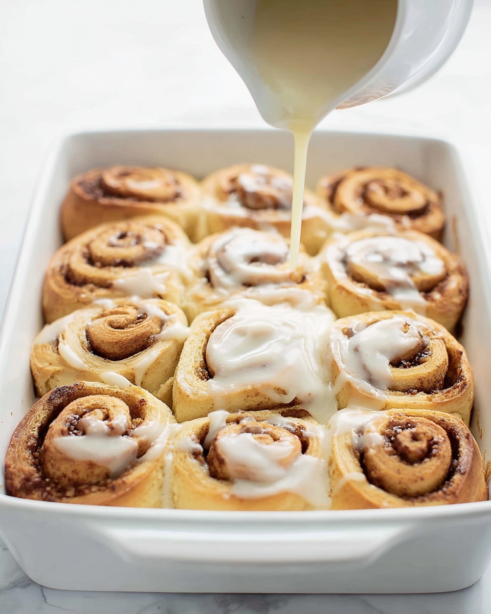 A white rectangular baking dish holds nine golden-brown cinnamon rolls arranged in a 3x3 grid, each roll showing spiral layers of soft dough and cinnamon filling with a slightly crispy outer edge. A pale cream sauce is being poured from a white jug at the top-center, slowly dripping onto the rolls and beginning to cover two rows at the bottom with a smooth, shiny layer of icing. The scene is set on a white marbled surface, creating a clean and bright background. Photo taken with an iphone --ar 4:5 --v 7