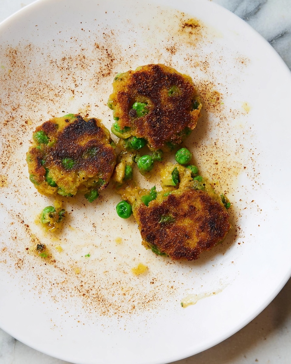 The image shows three small, round, golden-brown patties with green peas visible inside, placed on a white plate. The patties have a slightly crispy texture with some parts darker brown from frying. The plate has some oil stains and light brown powder sprinkled around. The background is a white marbled surface. photo taken with an iphone --ar 4:5 --v 7