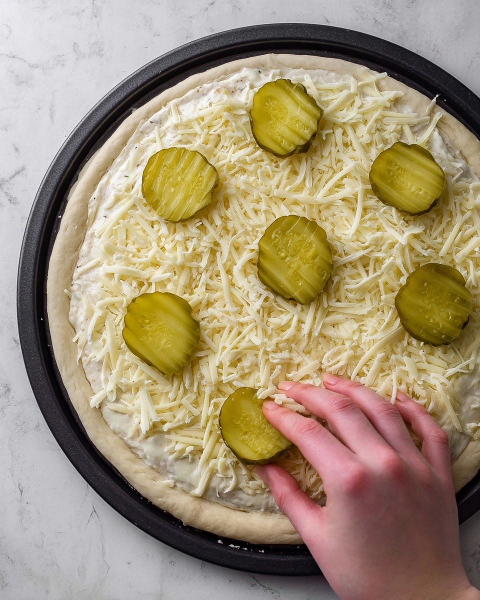 The image shows a white pizza dough spread on a round black pizza pan placed on a white marbled surface. The dough is topped evenly with a layer of white sauce, followed by a layer of shredded white cheese that covers almost all the surface inside the crust. On top of the cheese, there are four green pickle slices placed in a loose diagonal line. A woman's hand is reaching from the right side to place the last pickle slice near the edge of the pan. Photo taken with an iphone --ar 4:5 --v 7