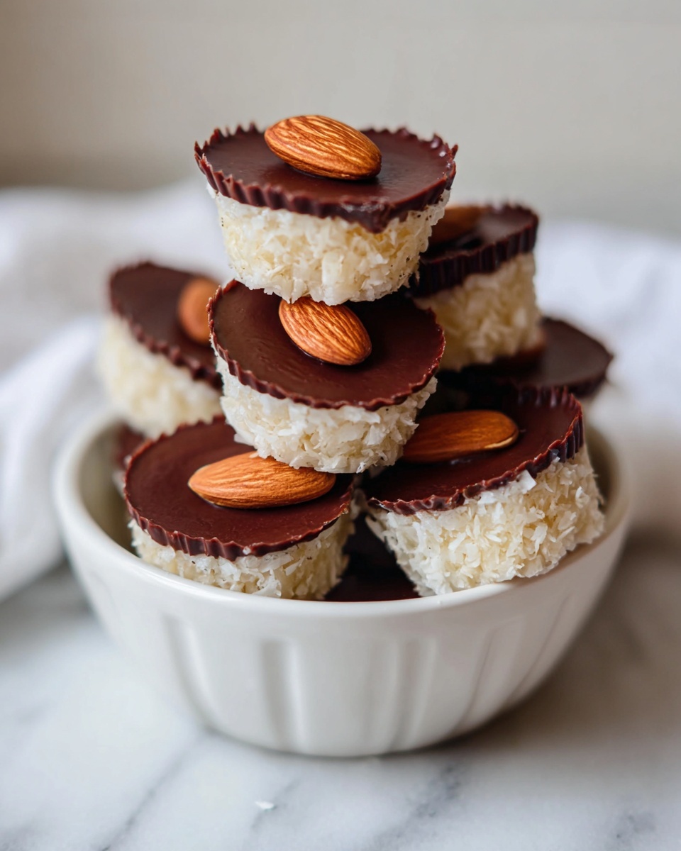 A small white bowl filled with bite-sized treats stacked in a loose pile, each treat has two visible layers: the bottom layer is a rough-textured, off-white base resembling shredded coconut, and the top layer is a smooth, dark brown chocolate disk with a single whole almond pressed into the center. The bowl sits on a white marbled surface with soft lighting that highlights the textures and colors of the treats, photo taken with an iphone --ar 4:5 --v 7