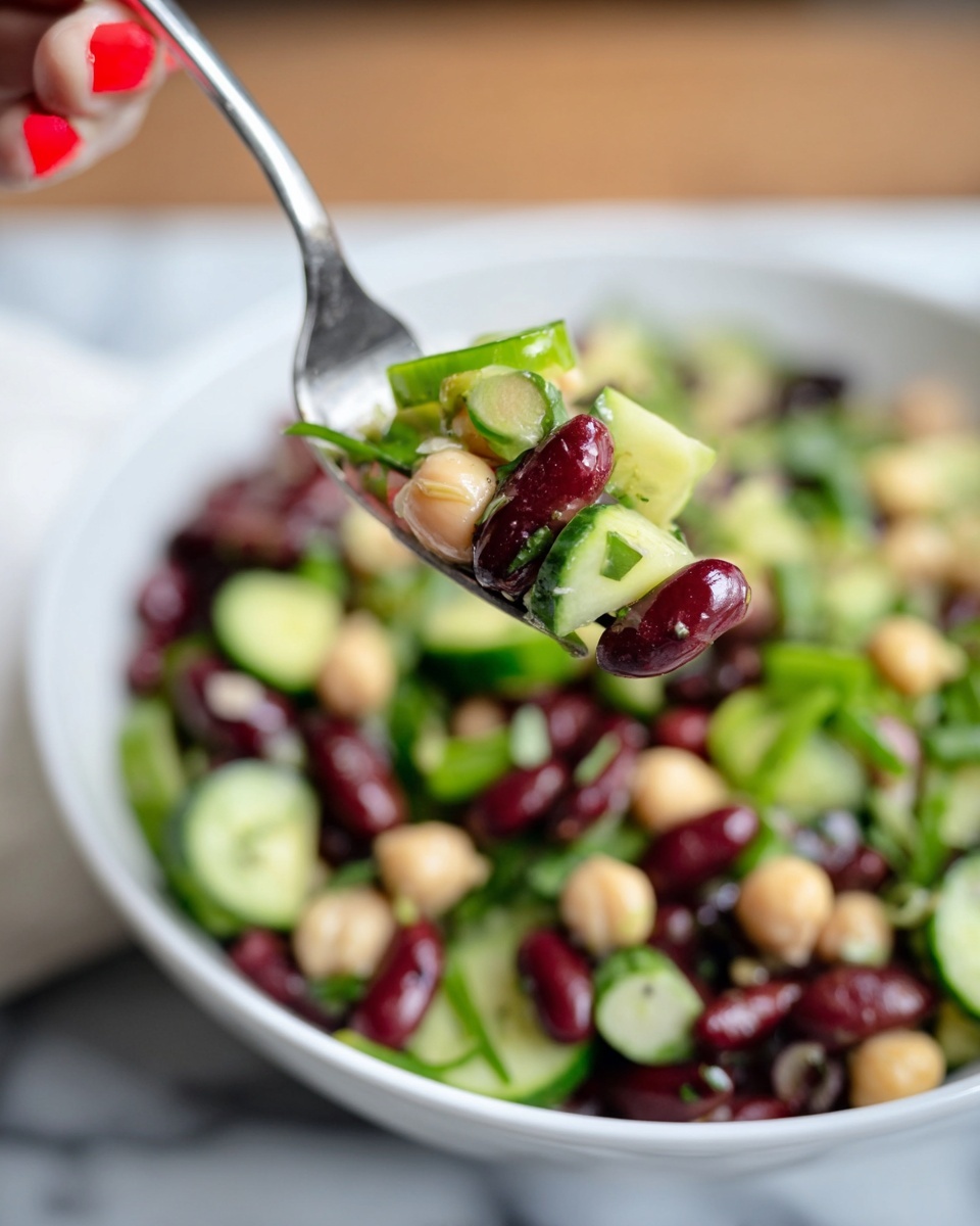 A white bowl filled with a colorful bean salad sits on a white marbled surface, showing at least four main layers: dark red kidney beans, light beige chickpeas, small black beans, and bright green sliced cucumbers. Thin green slices of scallions or herbs are mixed throughout, adding a fresh texture. The beans have a smooth and shiny surface, while the cucumber slices have a fresh, slightly translucent look with a light green peel. In the foreground, there is a close-up of a shiny fork lifting a bite with kidney beans, cucumber slices, and herbs, held by a woman's hand with red-painted nails. The background is softly blurred, focusing on the salad’s vibrant colors and the fork’s contents. photo taken with an iphone --ar 4:5 --v 7