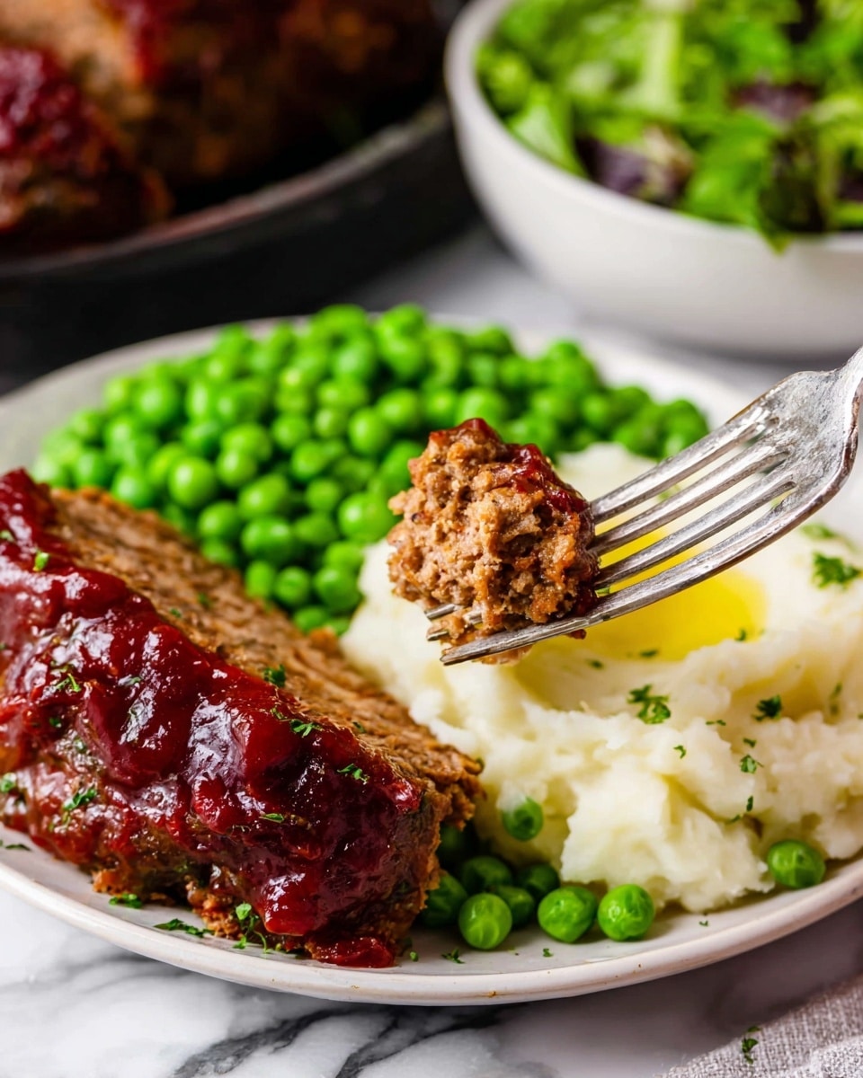 A white plate holds a meal with three main layers: bright green peas with a fresh texture on the left, creamy mashed potatoes with a smooth texture and a small pool of melted butter in the middle, and a thick slice of brown meatloaf with a shiny, dark red sauce on top on the right. A fork in the foreground lifts a small piece of the meatloaf showing a juicy, grainy texture with bits of sauce. The background shows more meatloaf and a bowl of leafy salad on a white marbled surface. The photo taken with an iphone --ar 4:5 --v 7