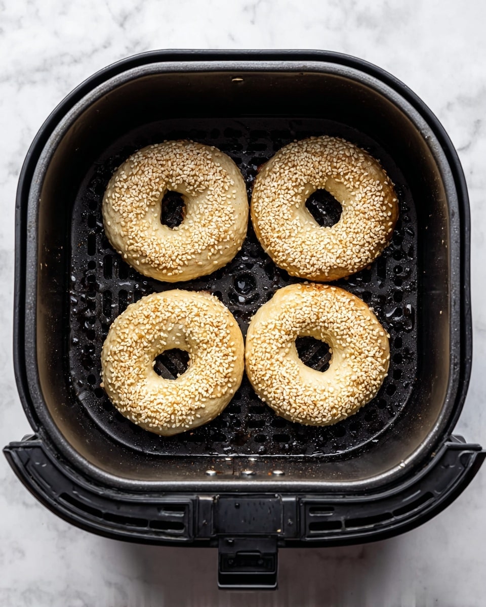 Inside a black air fryer basket with a perforated bottom, four round bagels are placed in a square shape. Each bagel is pale beige in color, topped with a scattering of white sesame seeds that add texture. The surface of the bagels looks soft and slightly puffy, while the hole in the middle of each is irregular in shape. The basket sits on a white marbled surface, and little drops of moisture are visible on the black basket's bottom and sides. photo taken with an iphone --ar 4:5 --v 7