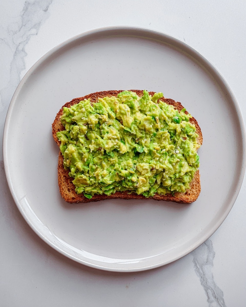 A single slice of toasted brown bread is placed in the center of a large white plate. On top of the toast is a thick, uneven layer of mashed avocado, showing a mix of light and dark green shades with small chunky bits creating a textured look. The toast edges are slightly darker and crisp, contrasting with the soft, creamy avocado spread. The plate rests on a white marbled surface, adding a subtle pattern in the background. photo taken with an iphone --ar 4:5 --v 7