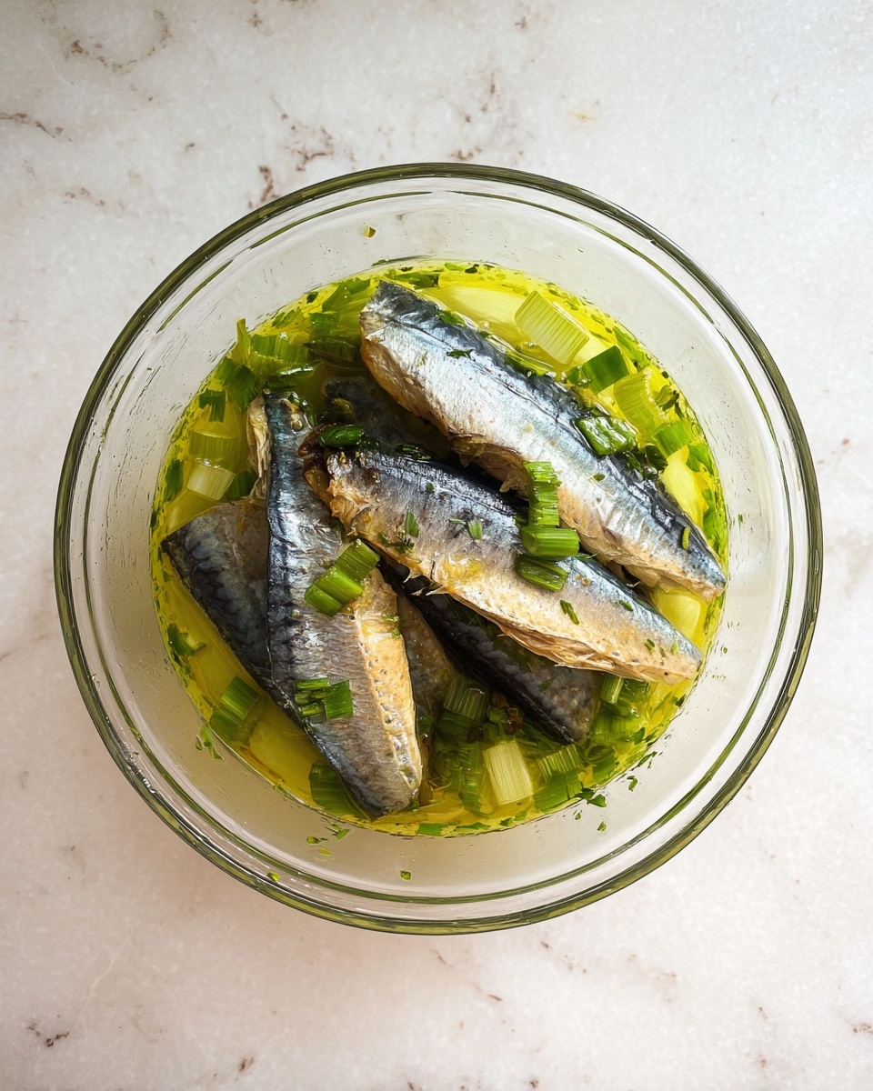 A clear glass bowl shows several fish pieces, with their silvery blue and brown skins facing up, stacked gently over each other. The fish sit in a greenish-yellow liquid with visible herbs and chopped green onions floating around, adding fresh green and yellow colors. The bowl is placed on a white marbled surface. photo taken with an iphone --ar 4:5 --v 7