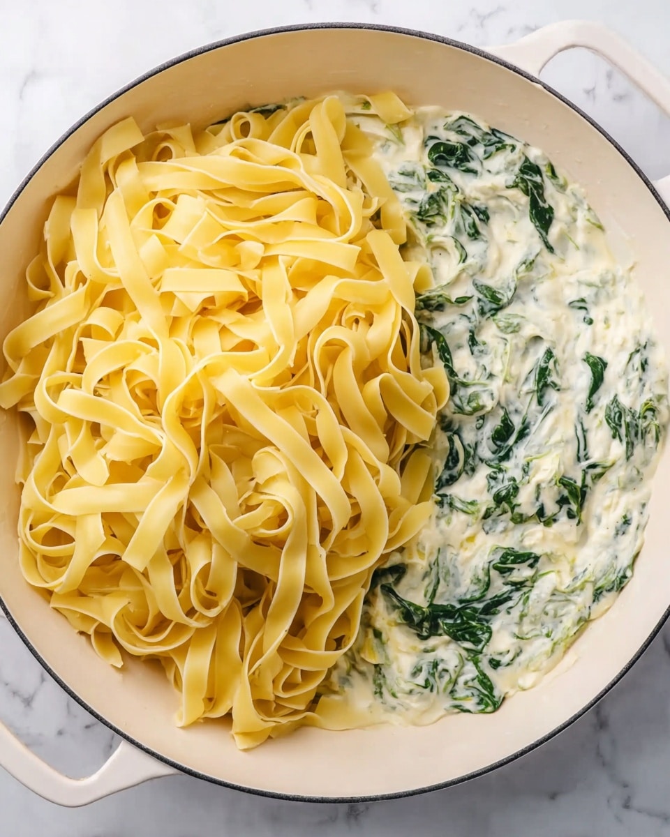 The image shows a white pan filled with two main layers. On the left side, there is a large pile of wide, flat yellow pasta noodles with a slightly shiny texture, loosely spread and overlapping each other. On the right side, the pasta sits partly in a creamy white sauce mixed with green spinach leaves, which adds a touch of bright green color and a soft, slightly wilted texture. The sauce looks rich and smooth, covering the bottom of the pan and blending into the greens. The pan is on a white marbled surface. photo taken with an iphone --ar 4:5 --v 7